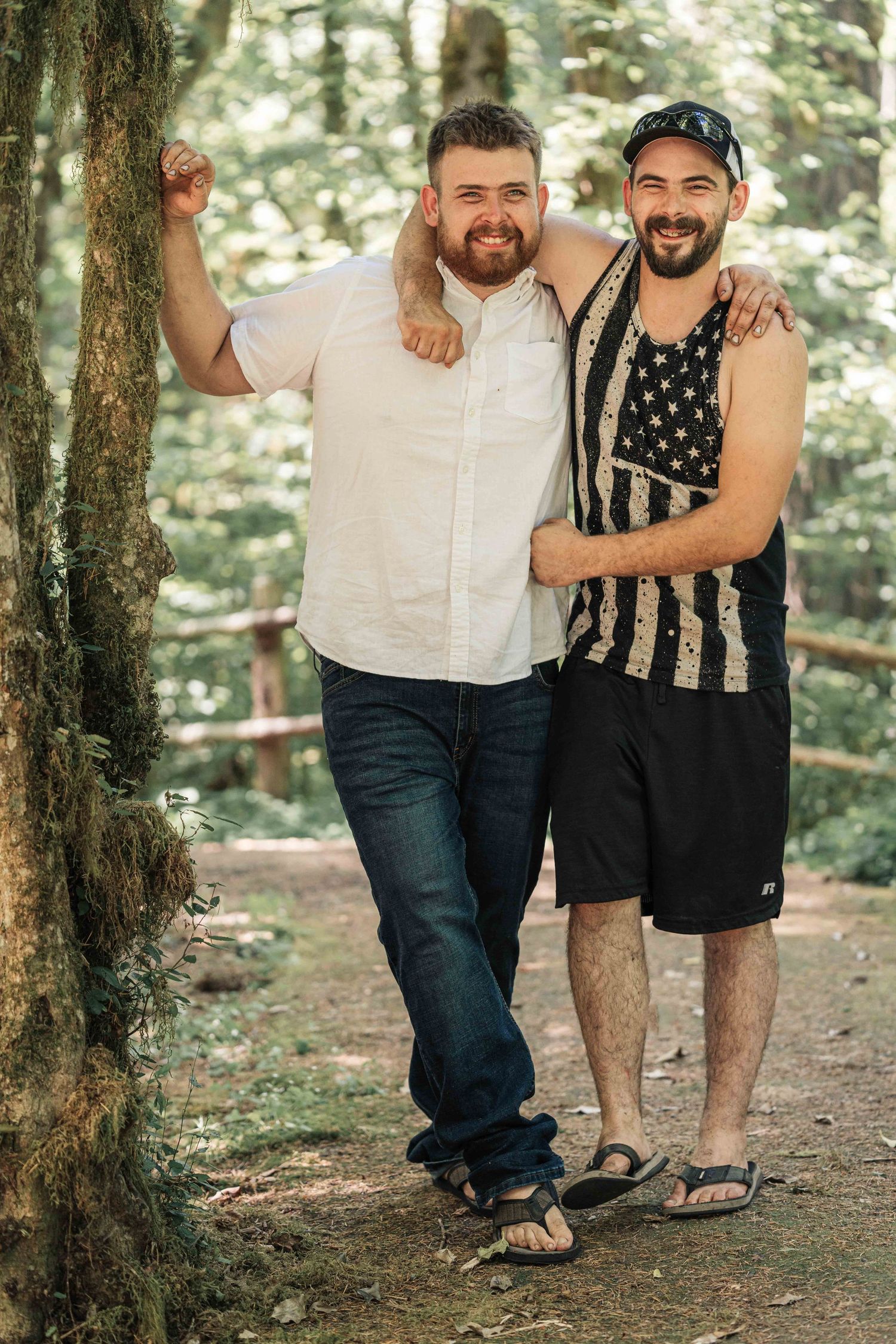 Two bearded men with arms around each other smile on a wooded trail, one in a white shirt and the other in an American flag tank top.