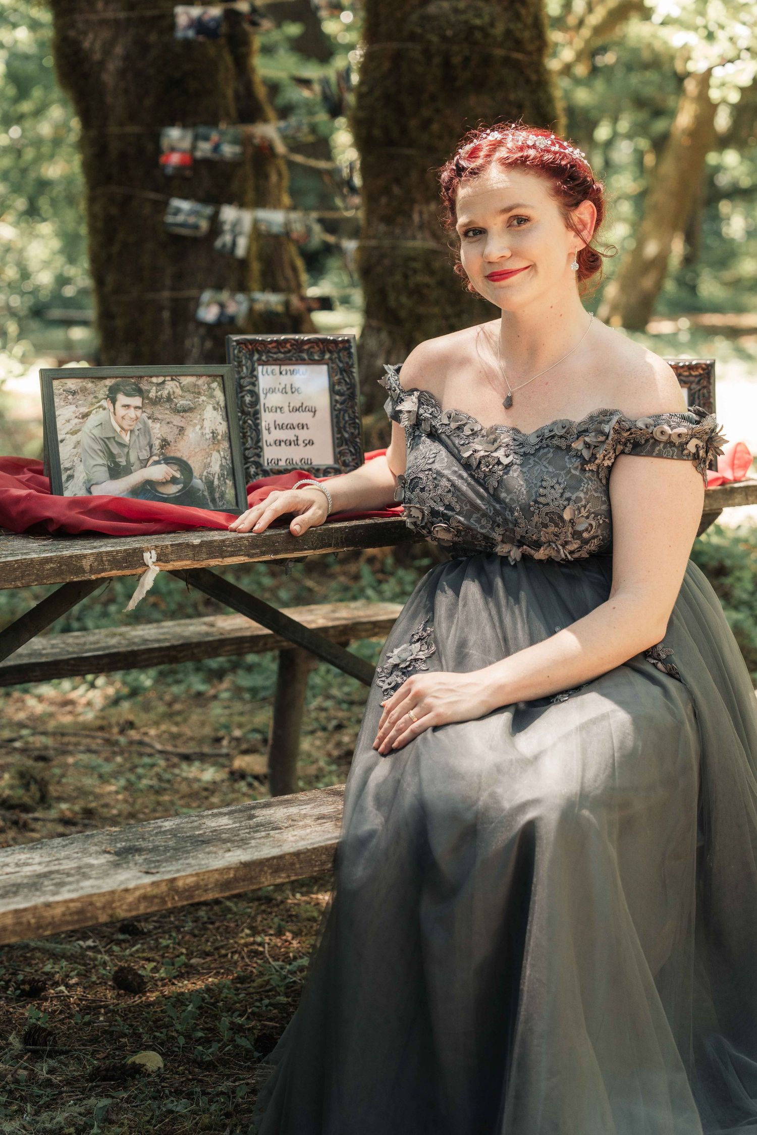 Smiling red-haired woman in a gray off-shoulder gown sits at a wooden bench beside framed photos at an outdoor forest ceremony.
