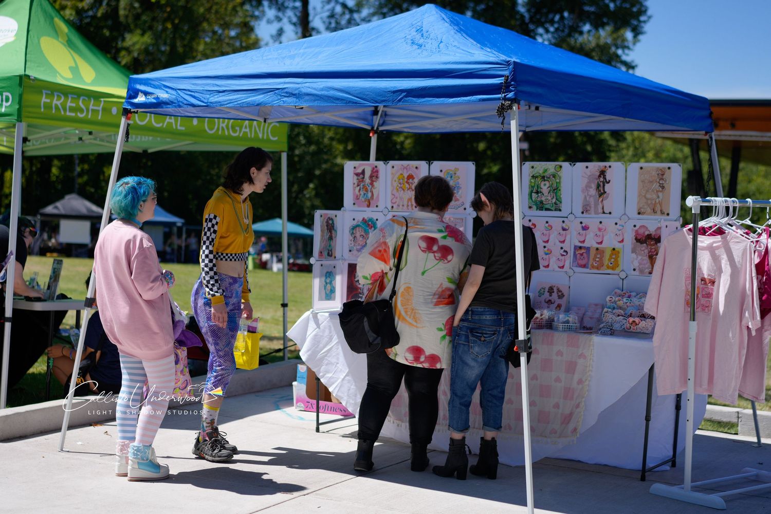 Visitors browse a vendor booth under a blue canopy tent displaying art prints and clothing at an outdoor community market.