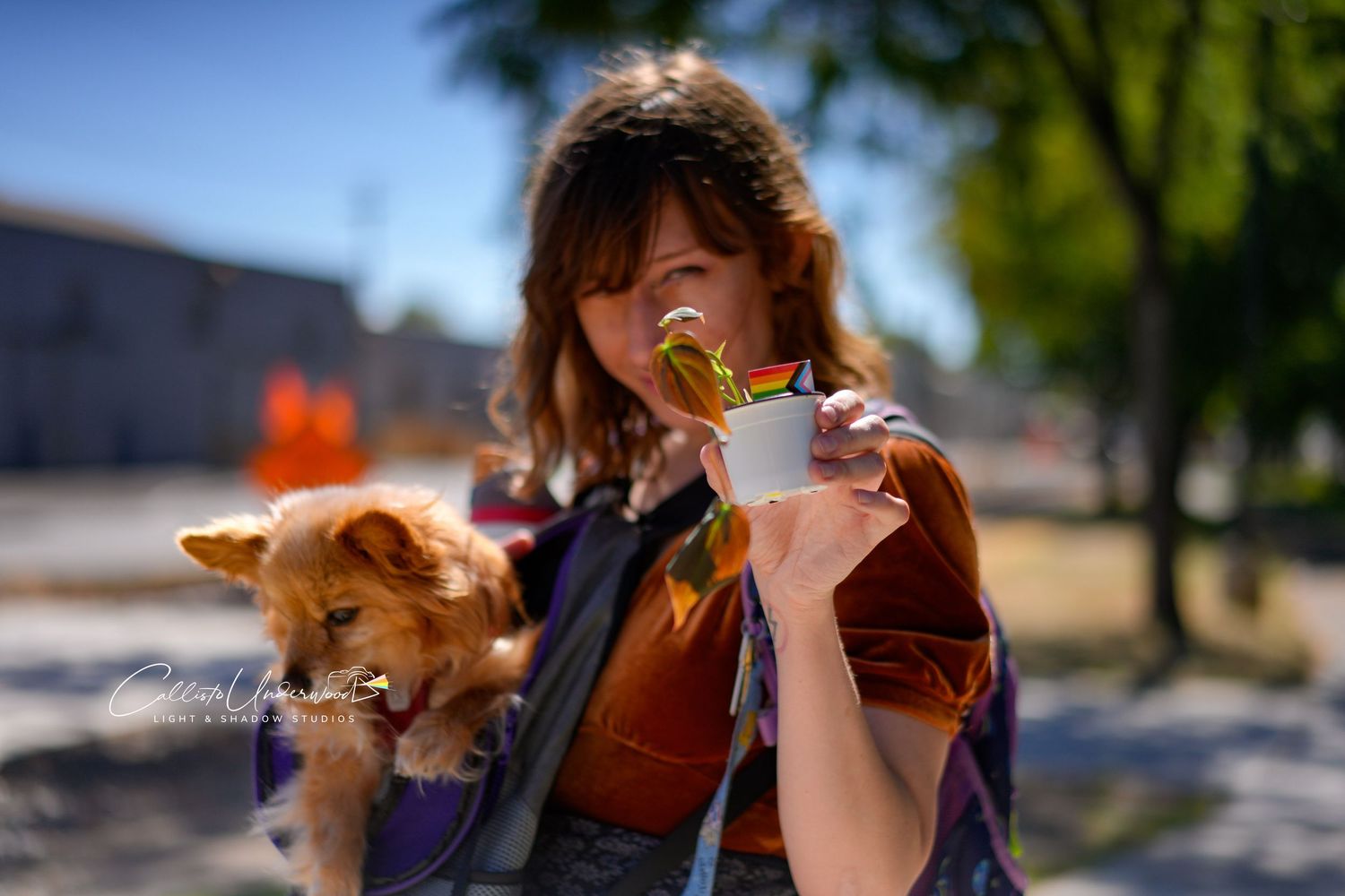 Smiling woman holds a fluffy tan dog under one arm and a small cup with a plant in the other hand on a sunny day outdoors.