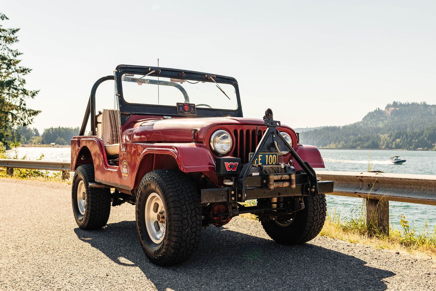 Vintage red Jeep CJ parked on a waterfront road with forested hills and calm water in the background.