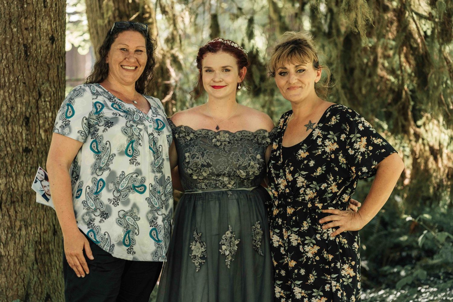 Three smiling women pose together among trees, one in a floral top, one in a gray tulle gown, and one in a black floral dress.