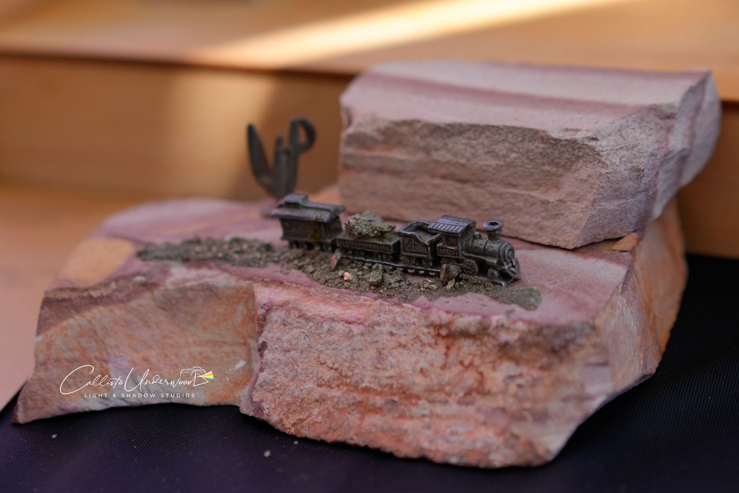Miniature metal steam locomotive model set on a piece of red sandstone with a tiny cactus, displayed at a craft fair.