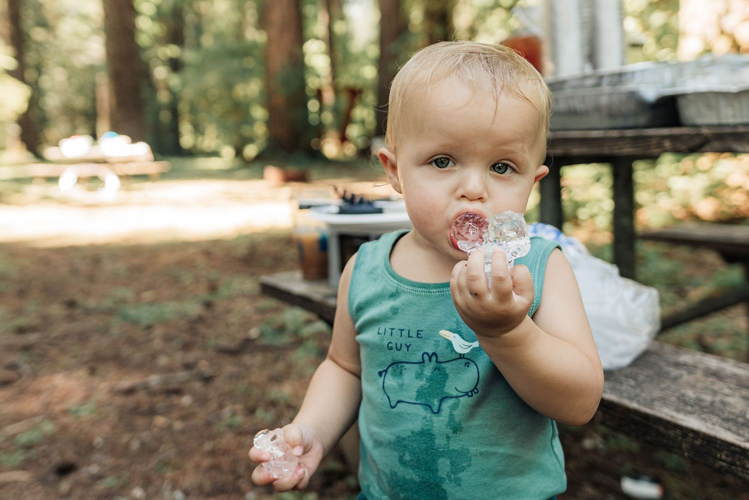 A toddler in a teal tank top sits at a wooden picnic table in the woods, eating a pink treat.
