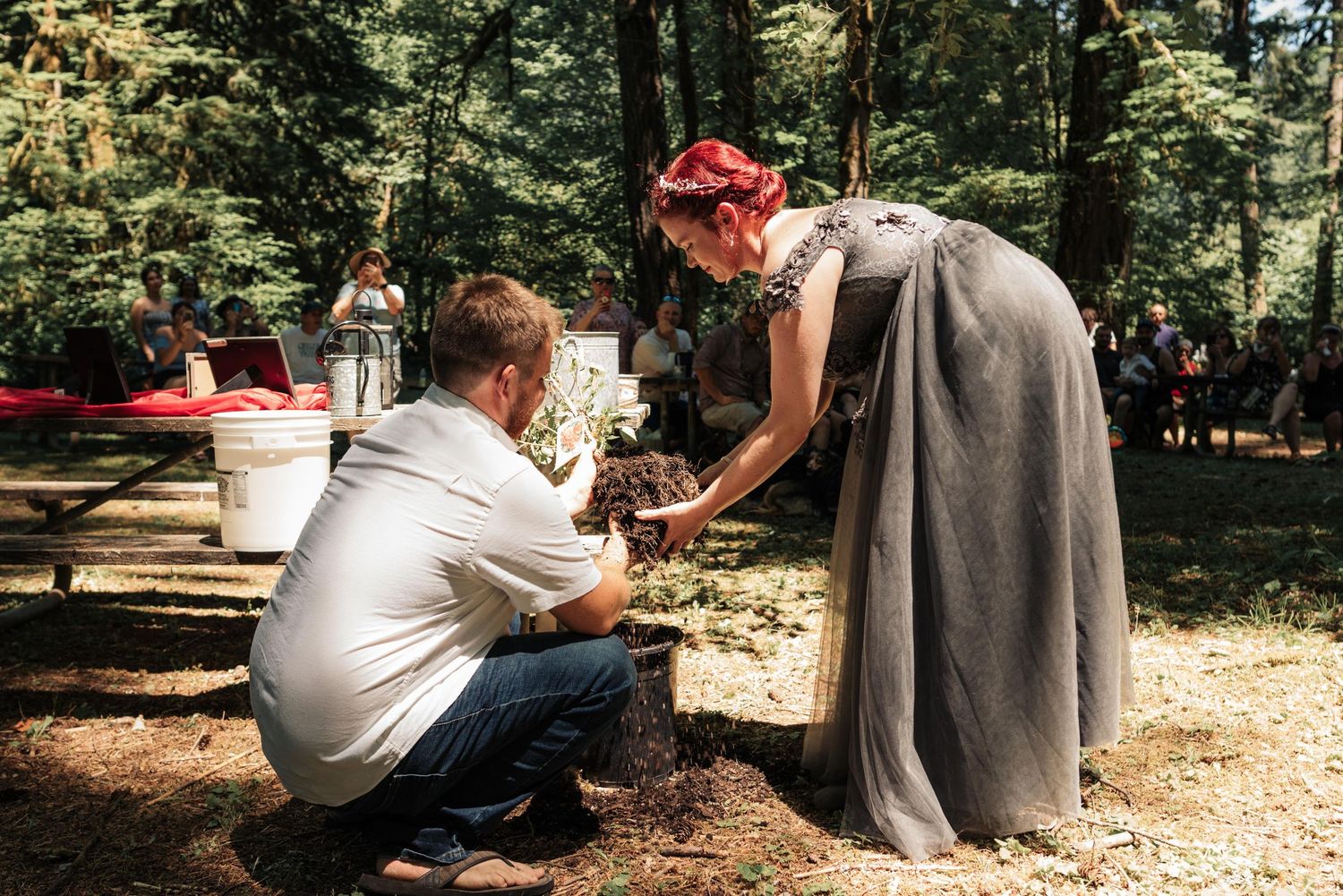 A bride in a gray gown and floral headpiece pours soil from her hands as a man in a white shirt kneels beside her at a forest wedding.