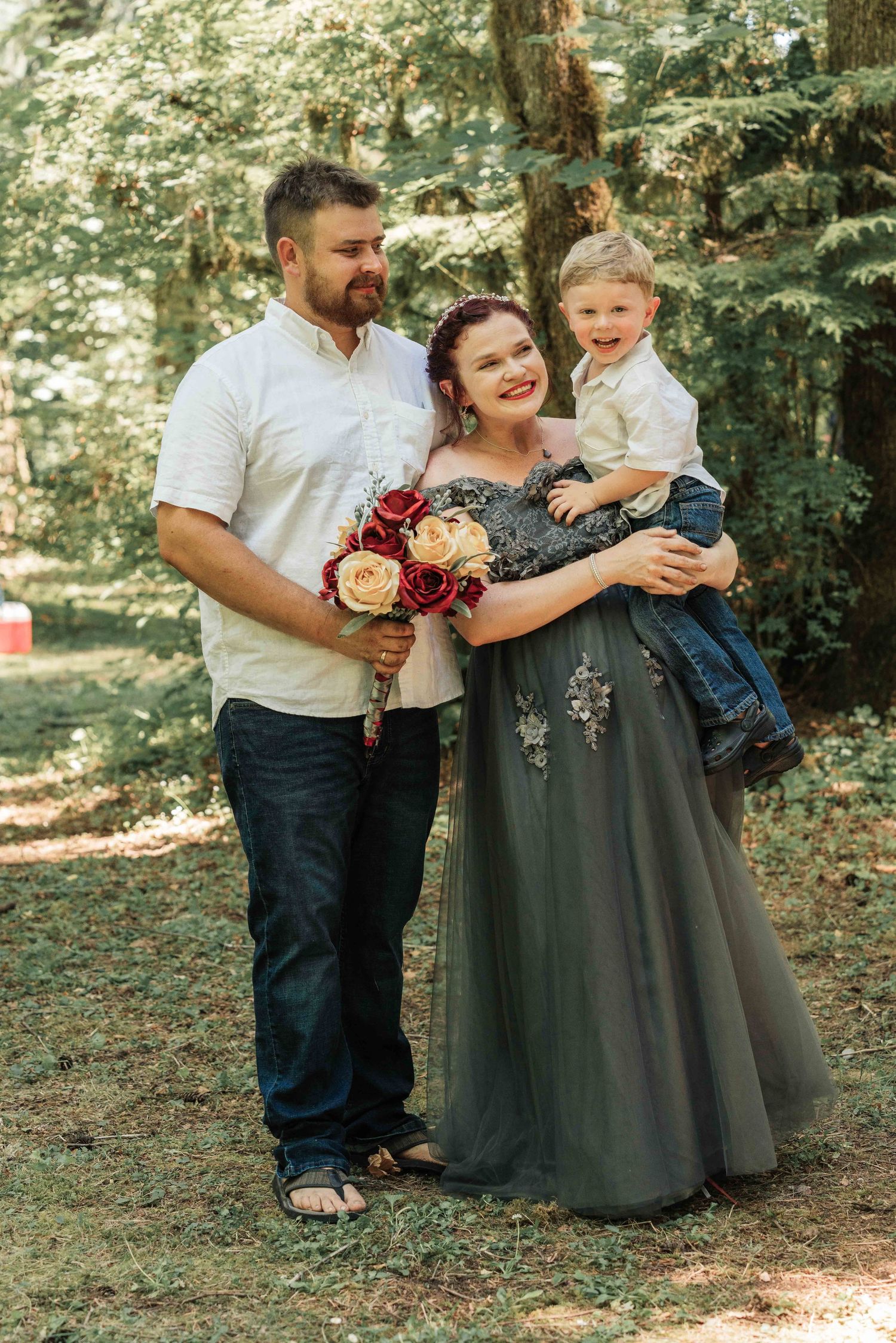 A happy family of three stands in a forest clearing, the bride in a gray gown holding her laughing son and a rose bouquet.