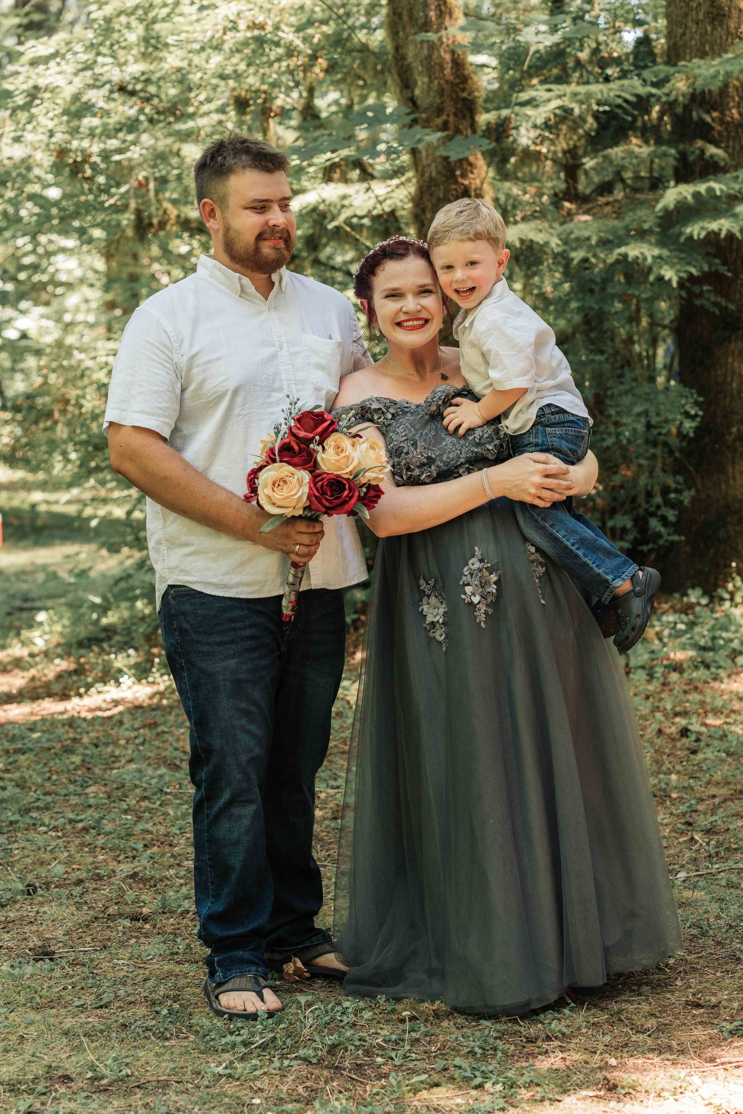 A smiling bride, groom and their young son pose together in a sunlit forest, the bride holding red and cream roses.