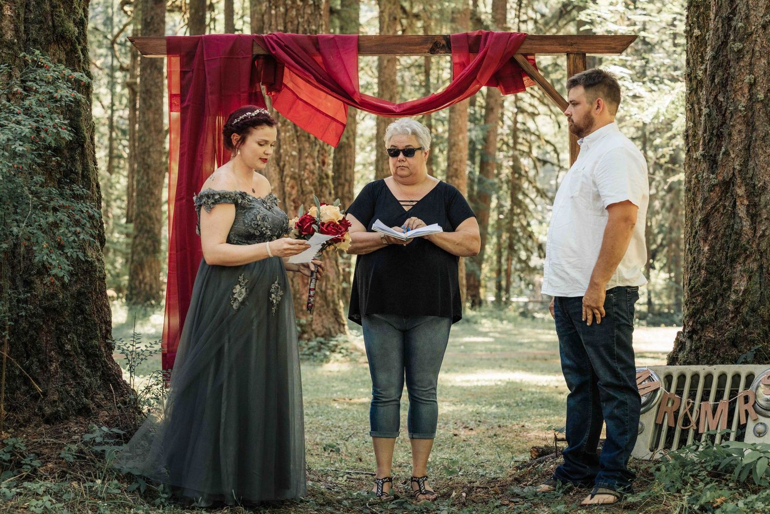 A bride holding a bouquet stands beside the officiant and groom during an outdoor forest wedding ceremony.