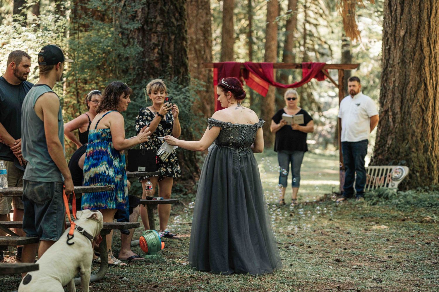 A bride in a gray off-the-shoulder gown stands before guests and a white dog at a forest wedding ceremony.