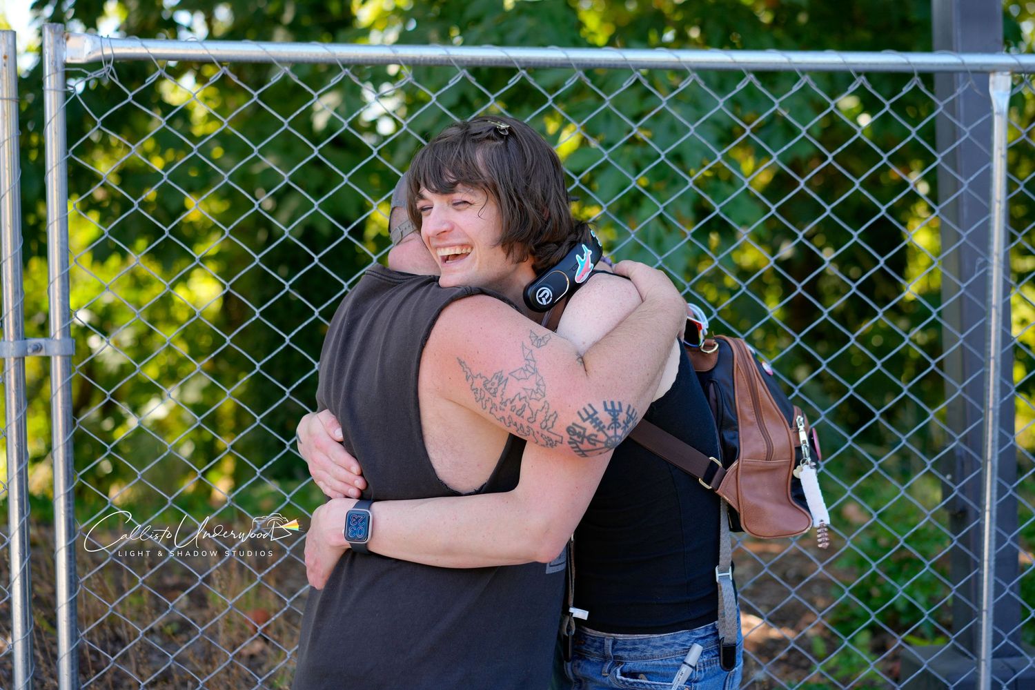 Two tattooed friends share a warm smiling hug in front of a chain-link fence with green foliage behind them.