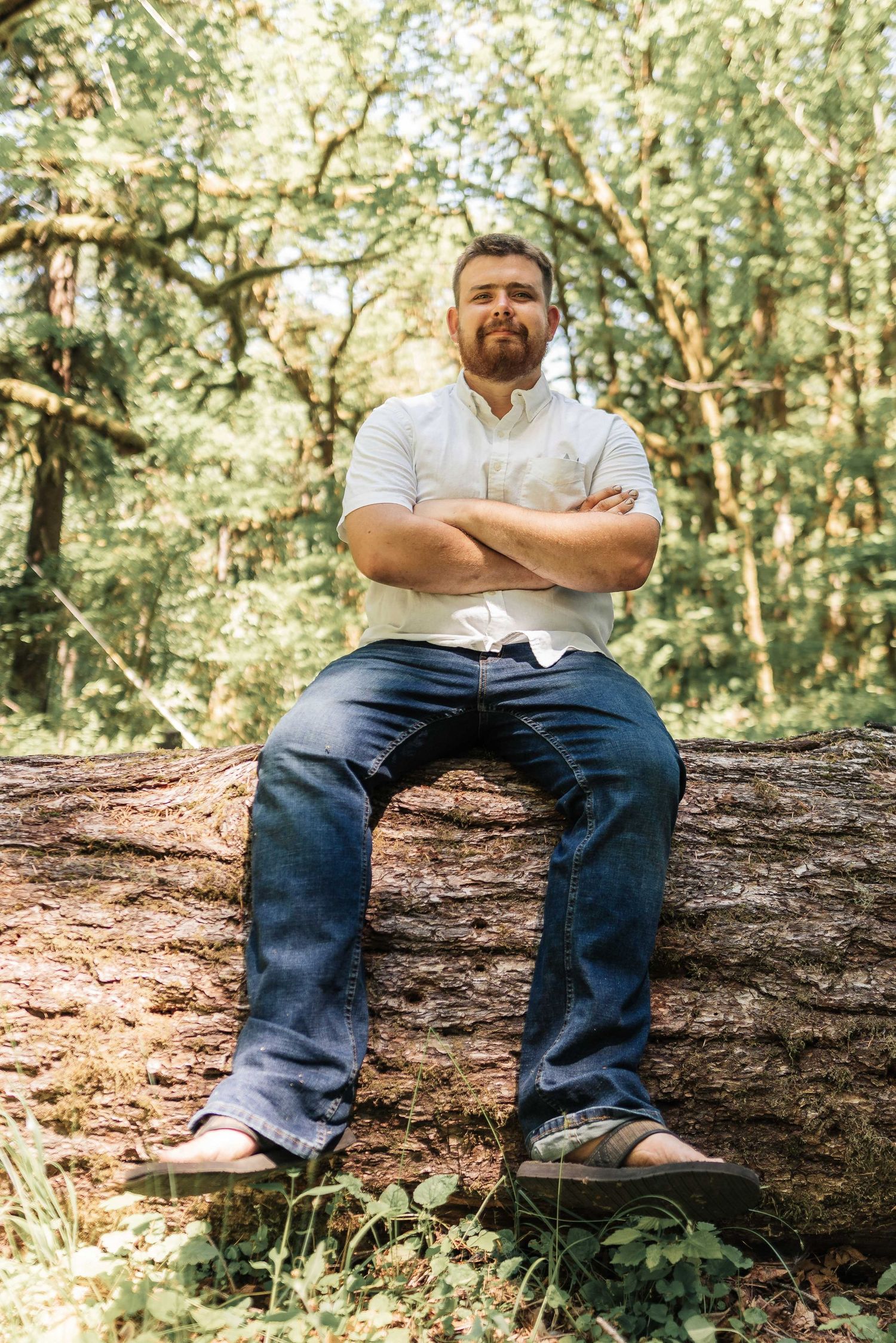 A bearded man in a white shirt and jeans sits with arms crossed on a large fallen log surrounded by green forest.