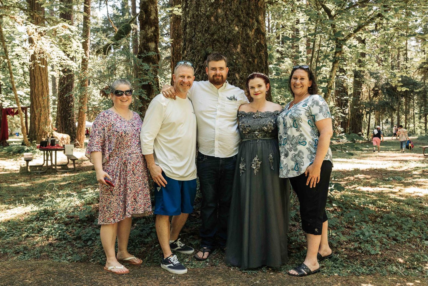 The bride and groom pose with four smiling family members in a sunlit forest clearing beneath tall evergreen trees.
