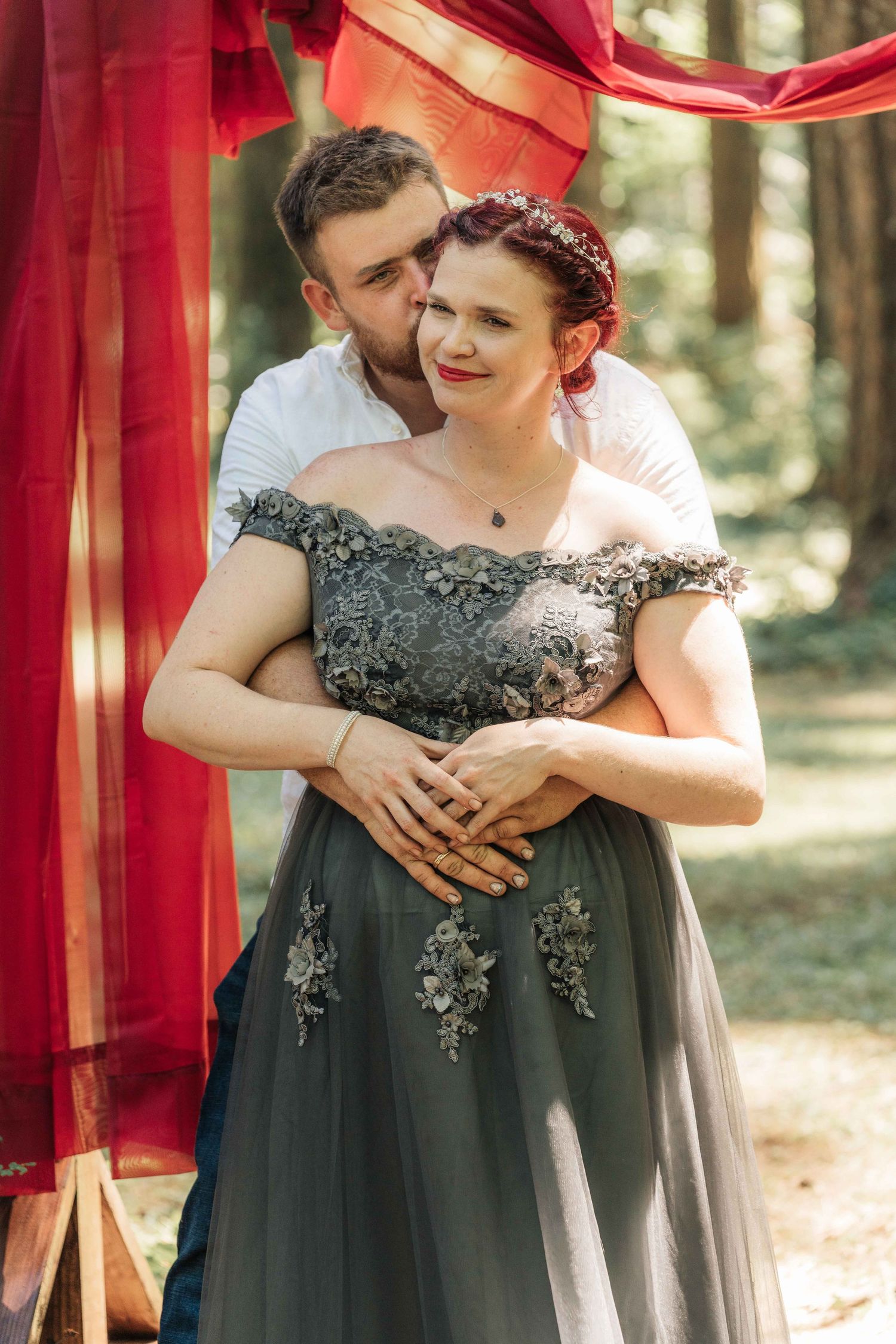 Man kisses the cheek of a smiling red-haired woman in a gray lace-detailed gown, framed by flowing red fabric in the woods.