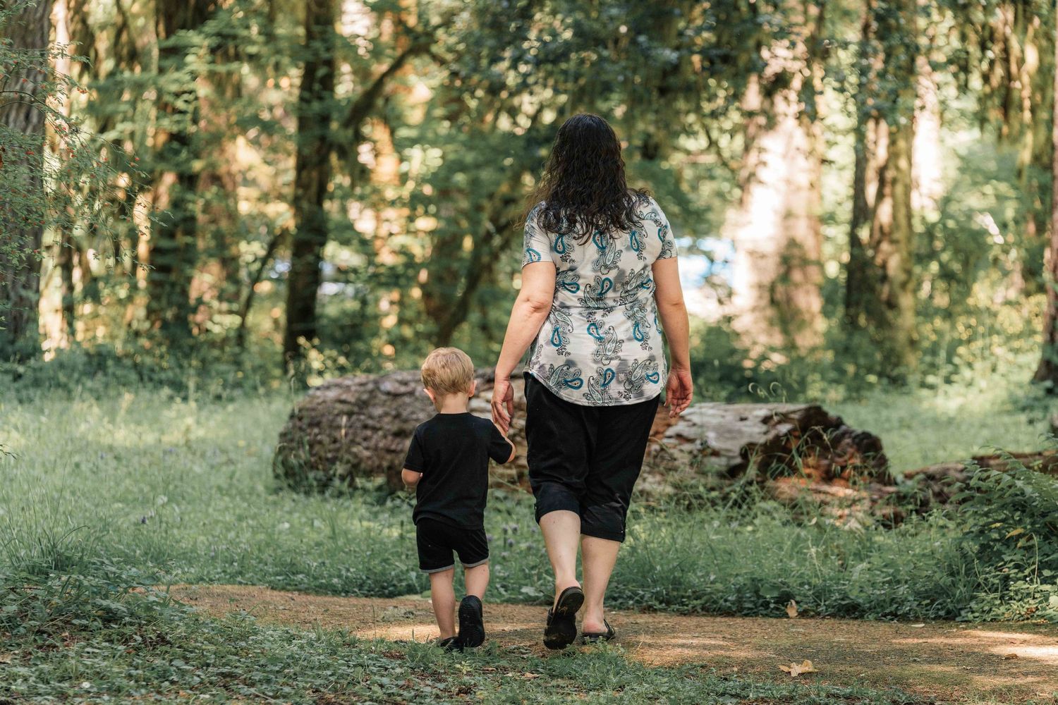 Woman in a floral top walks hand in hand with a toddler along a dirt path through a sunlit green forest.