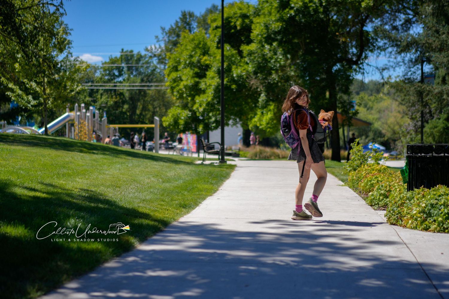 Woman with a backpack walks along a shaded park sidewalk bordered by green grass, shrubs and tall trees under a clear blue sky.