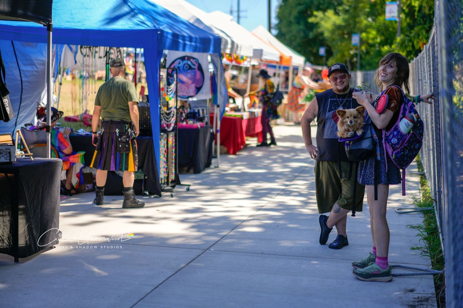 Festival street lined with blue canopy vendor tents as shoppers browse booths and a woman with a backpack talks to a man.