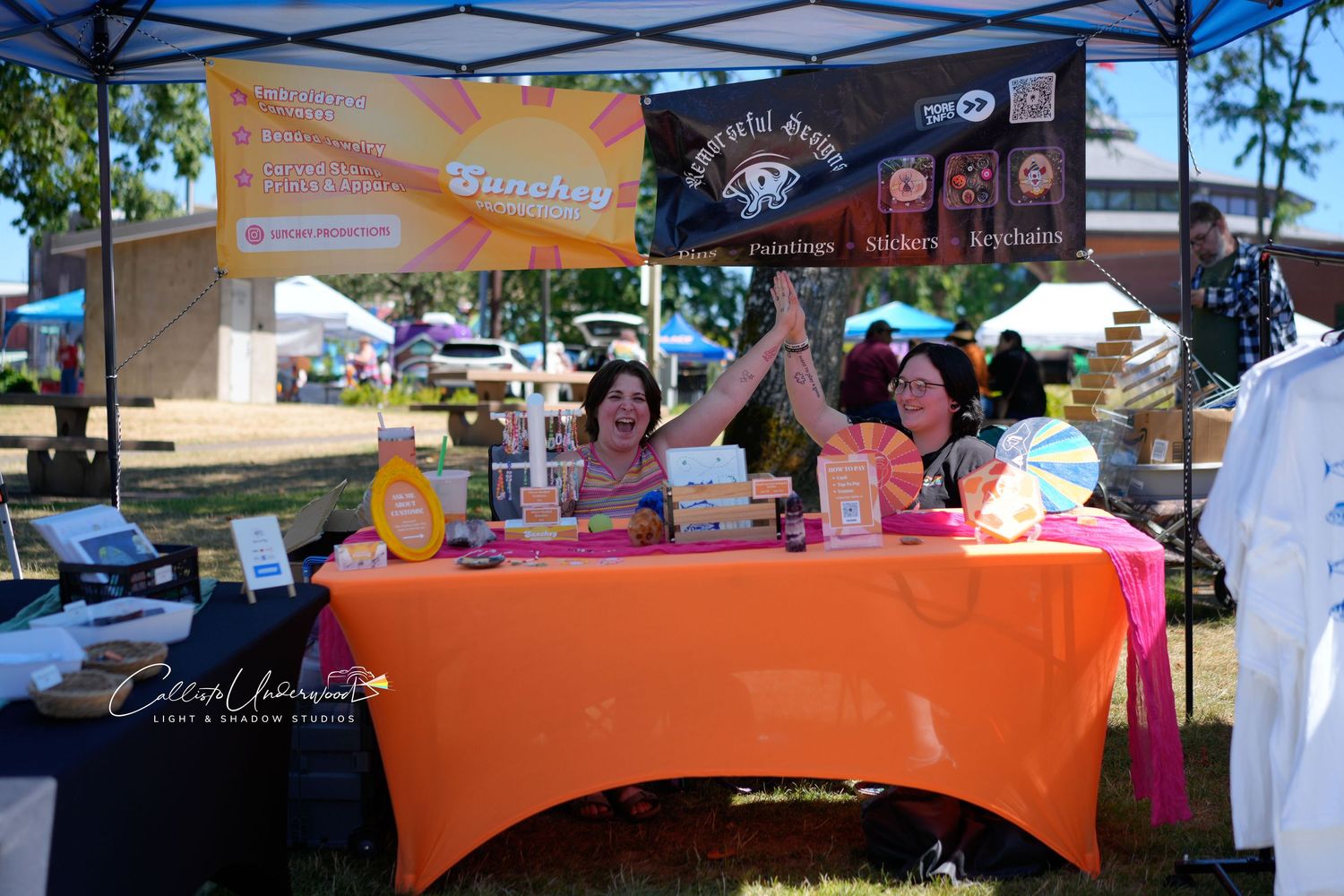 Two vendors raise their arms in celebration at an orange-covered market booth under a canopy with colorful banners at an outdoor fair.