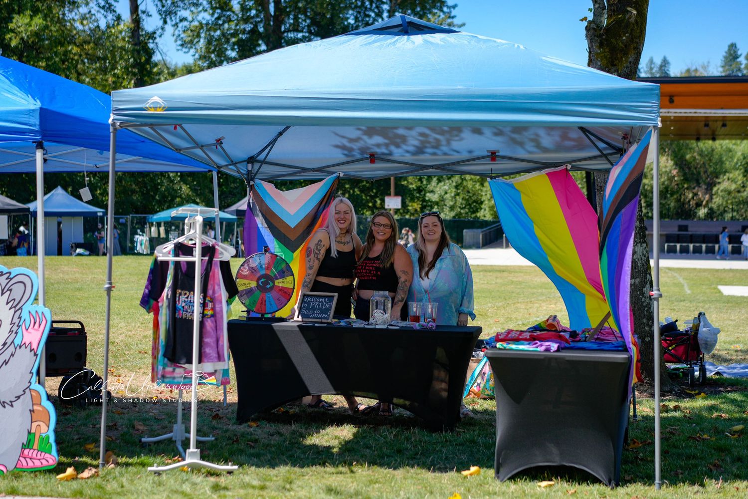 Three women smile behind a table under a light blue canopy tent at an outdoor craft fair, with rainbow fabric banners and hanging garments n