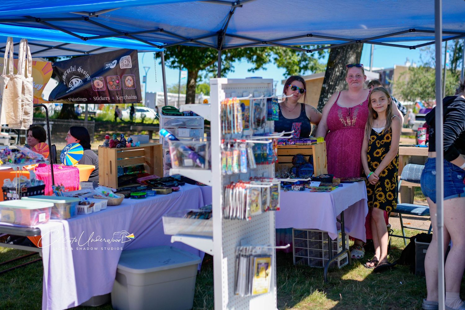 Three smiling vendors stand behind a craft booth with handmade goods and display racks at a sunny outdoor festival market.