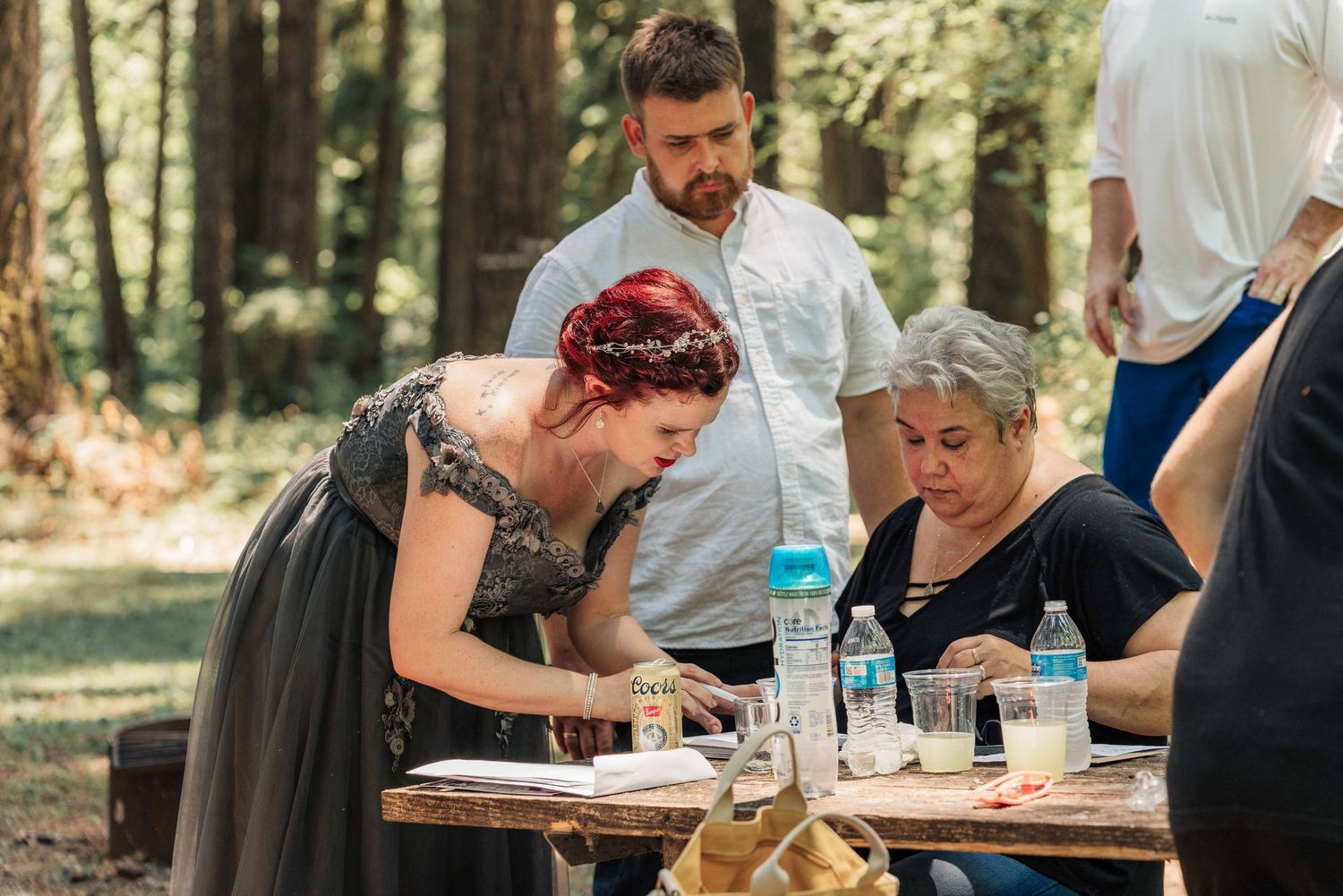 Woman in a gray dress with red hair writes at a wooden picnic table while a bearded man and an older woman look on in the woods.