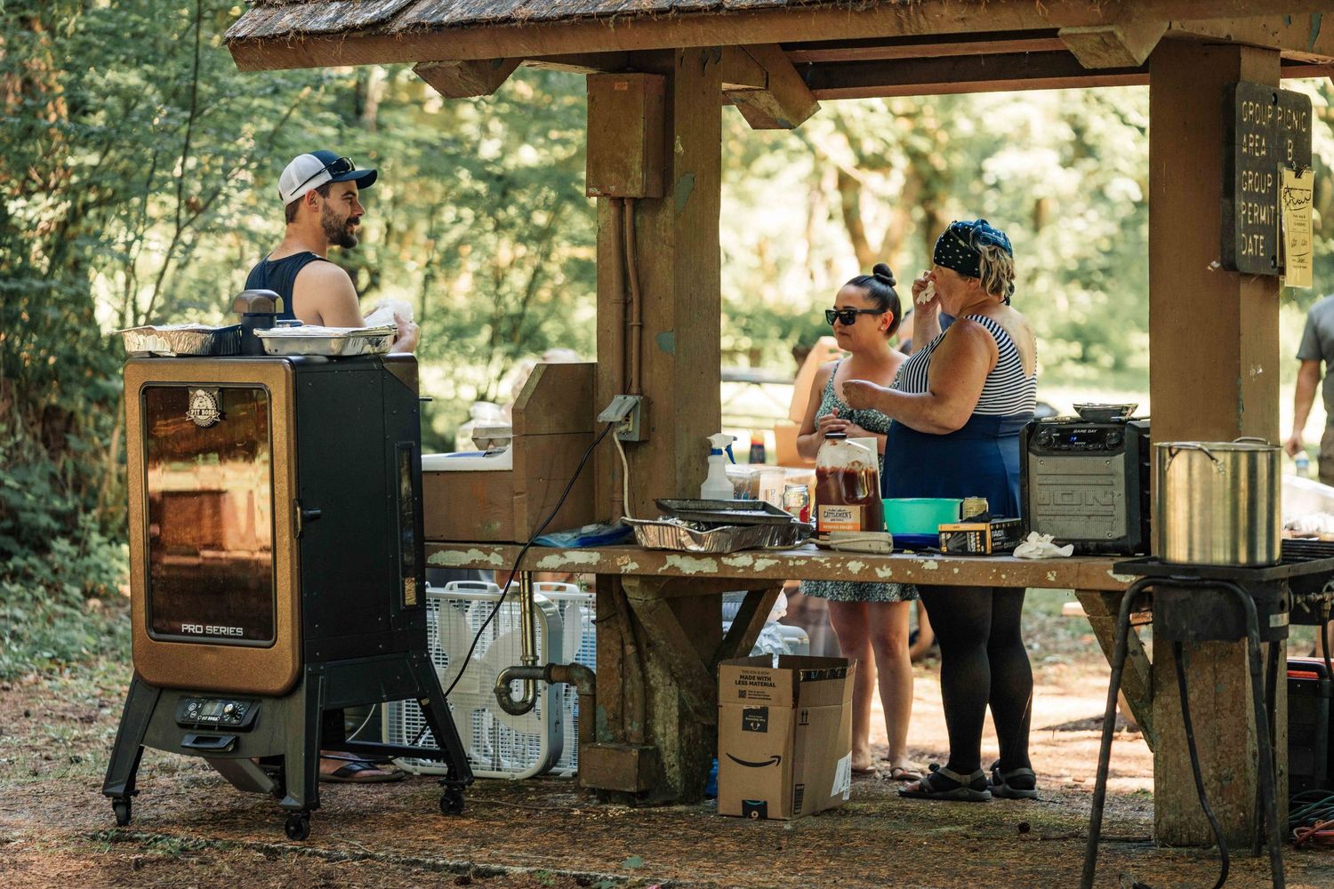 Guests prepare food at a rustic wooden outdoor pavilion with a vertical smoker and buffet table in the woods.