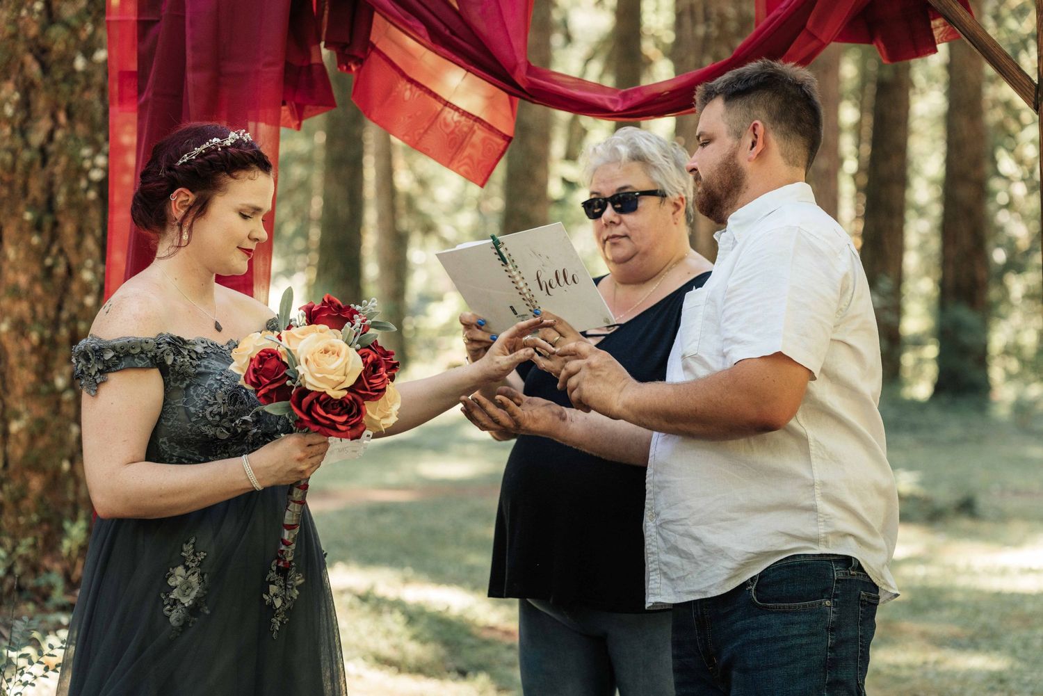 A couple holds hands while an officiant reads from a book during their outdoor forest wedding, the bride holding a rose bouquet.