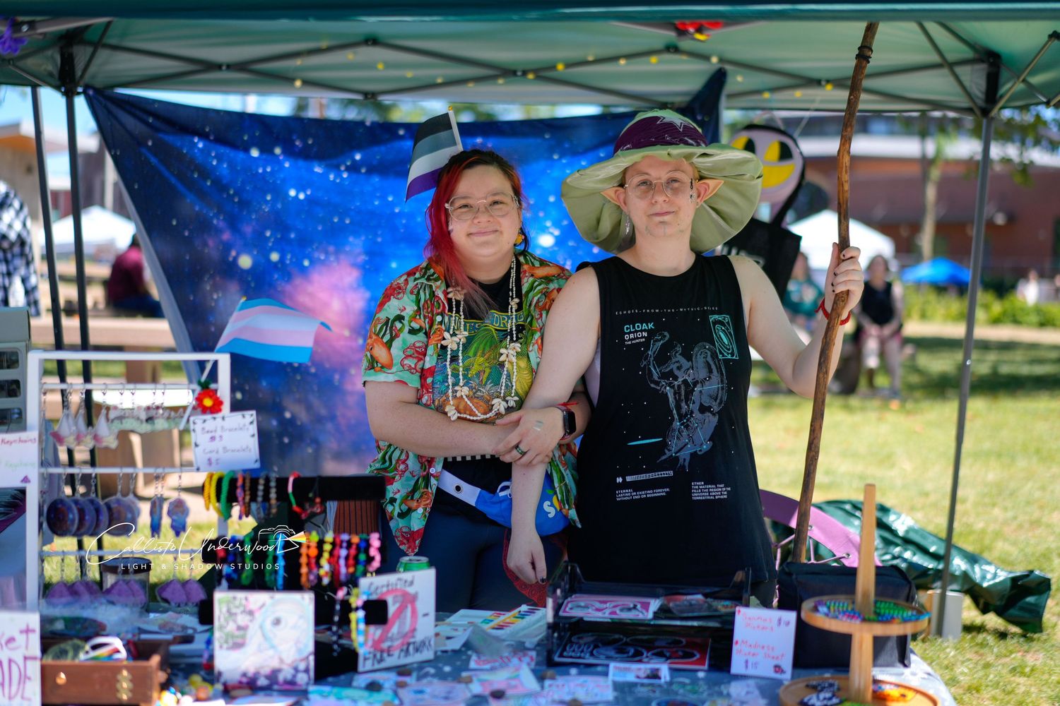 Two smiling vendors in colorful hats and clothing pose at a craft booth with jewelry and handmade items at an outdoor festival.