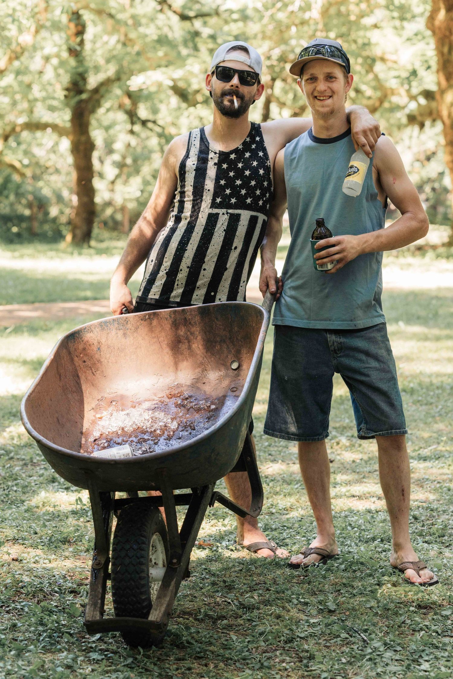 Two smiling men in caps pose beside a rusty wheelbarrow filled with ashes on a grassy lawn under tall trees.