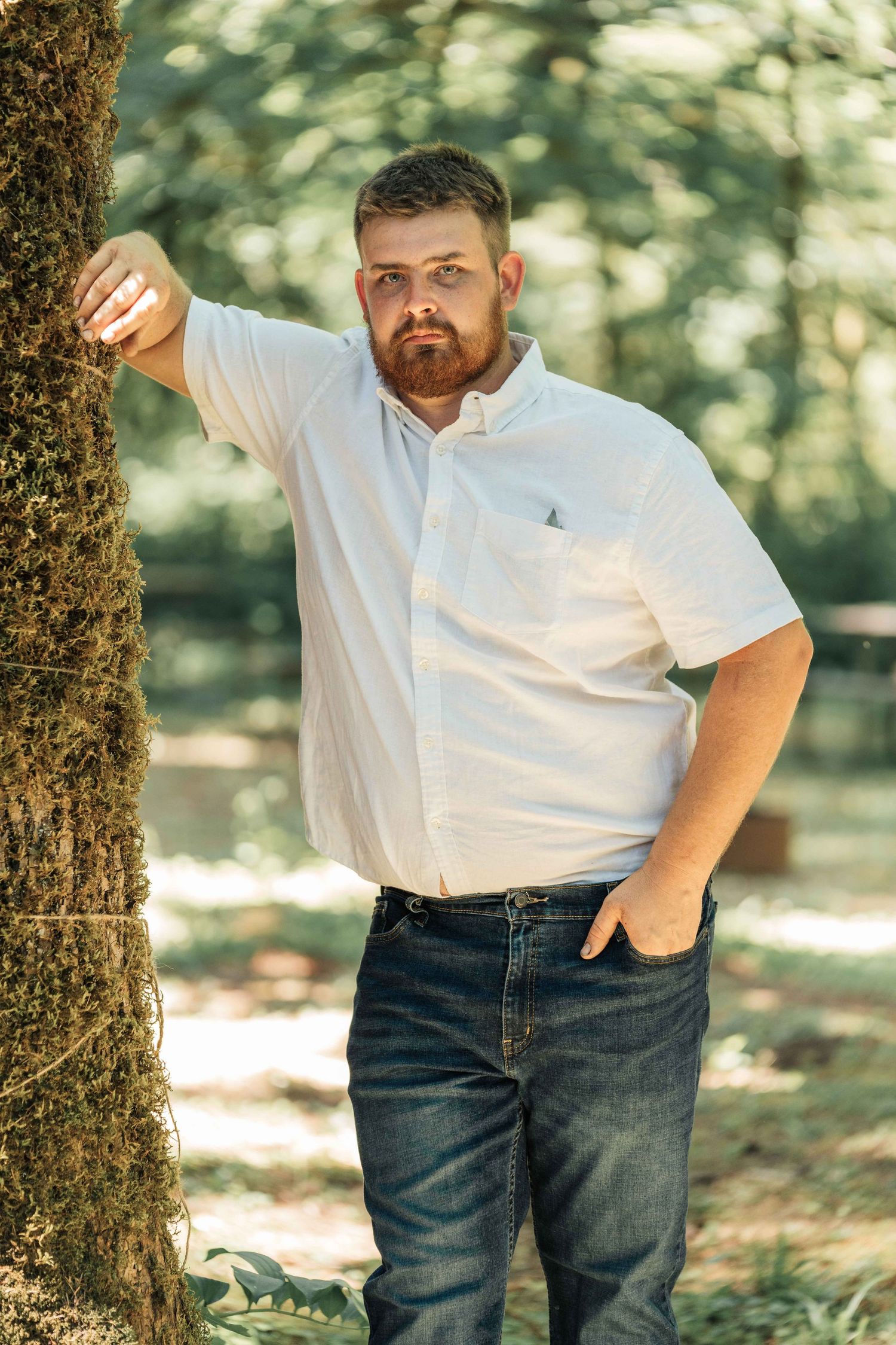 A bearded man in a white button-up shirt and jeans leans casually against a moss-covered tree in a sunlit forest.