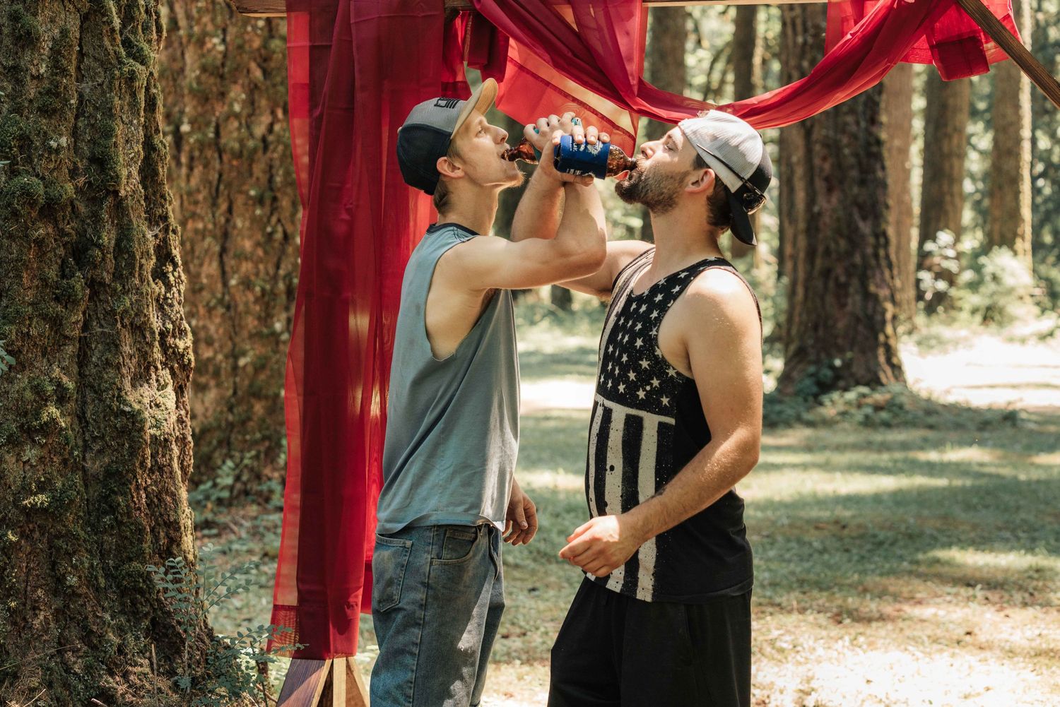 Two men in caps drink from water bottles under red draped fabric among tall trees during an outdoor gathering.