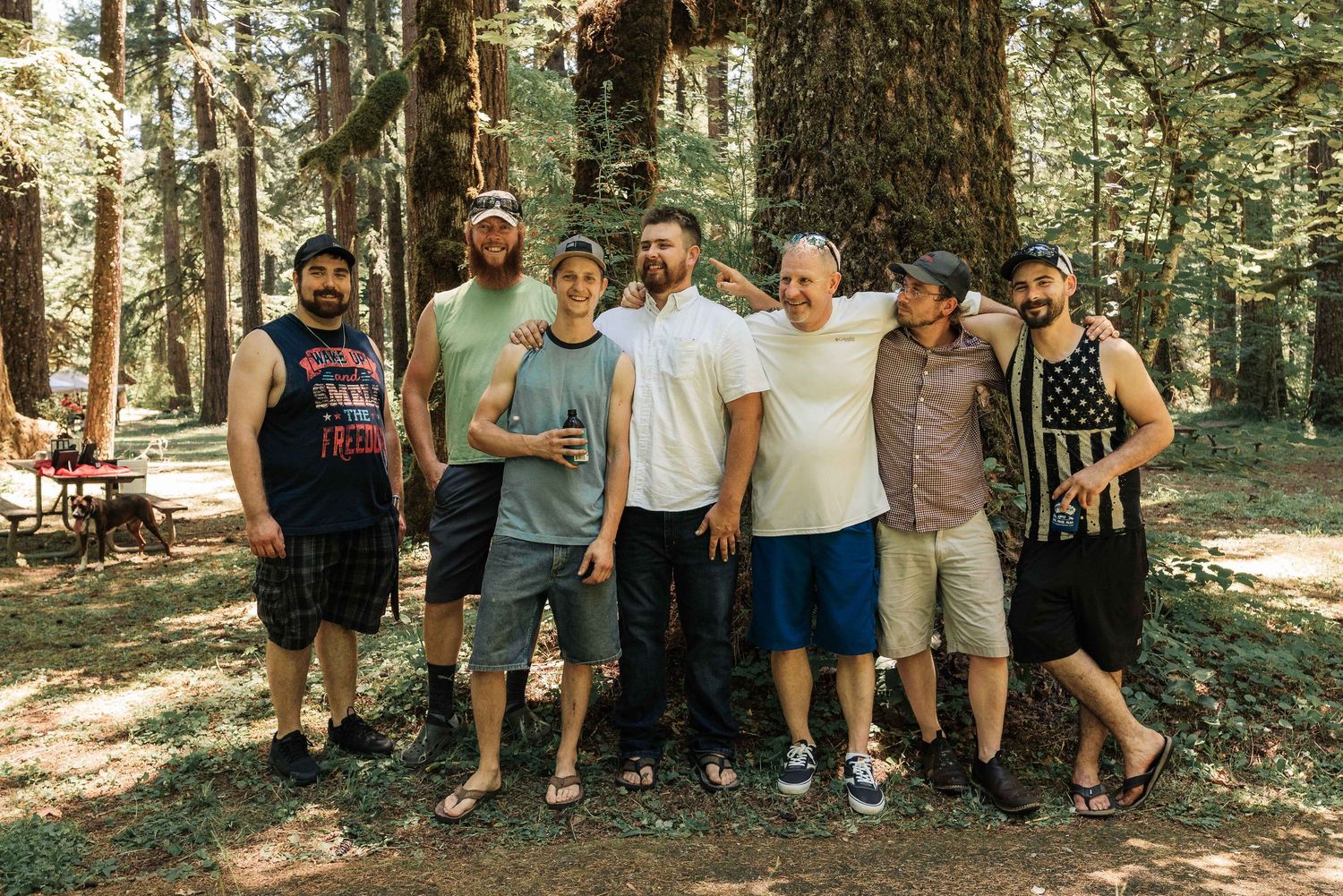 The groom and six smiling friends pose together in a sunlit forest, some holding drinks, with picnic tables in the background.