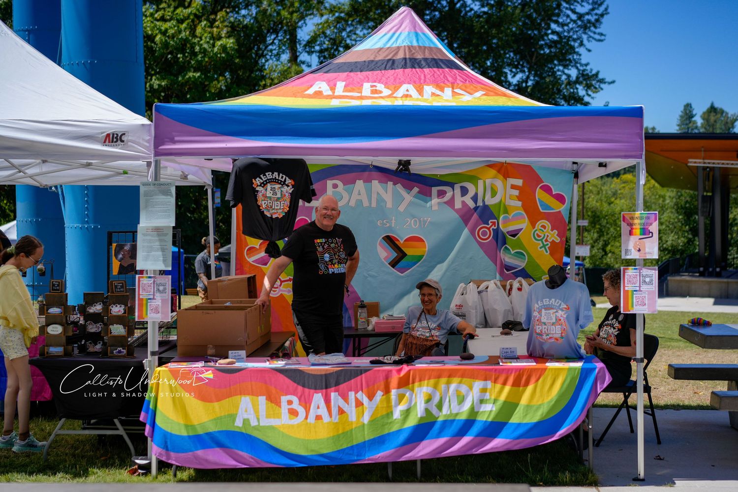 Colorful rainbow-draped Albany Pride booth with banner and volunteers under a canopy tent at an outdoor community event.