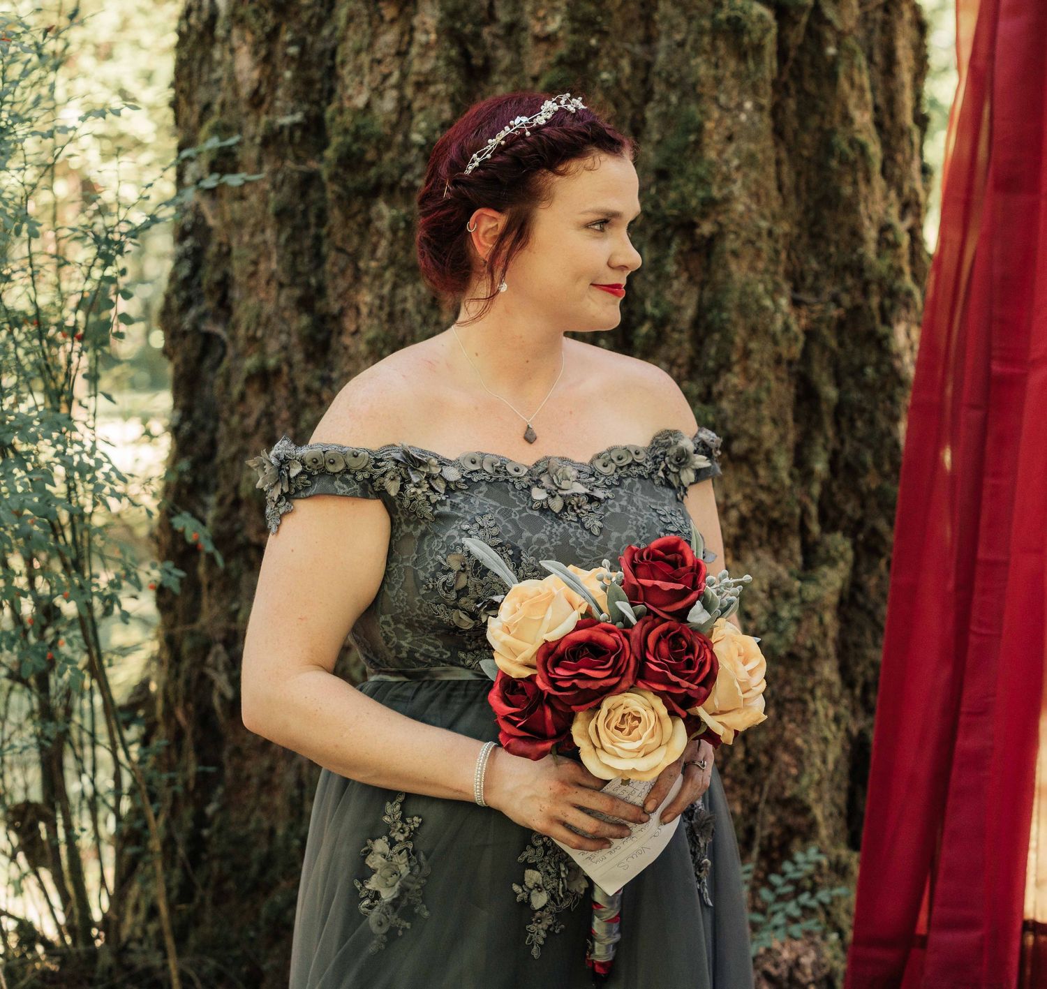 A bride with a floral hair crown and an off-shoulder gray lace gown holds a bouquet of red and cream roses beside a mossy tree.