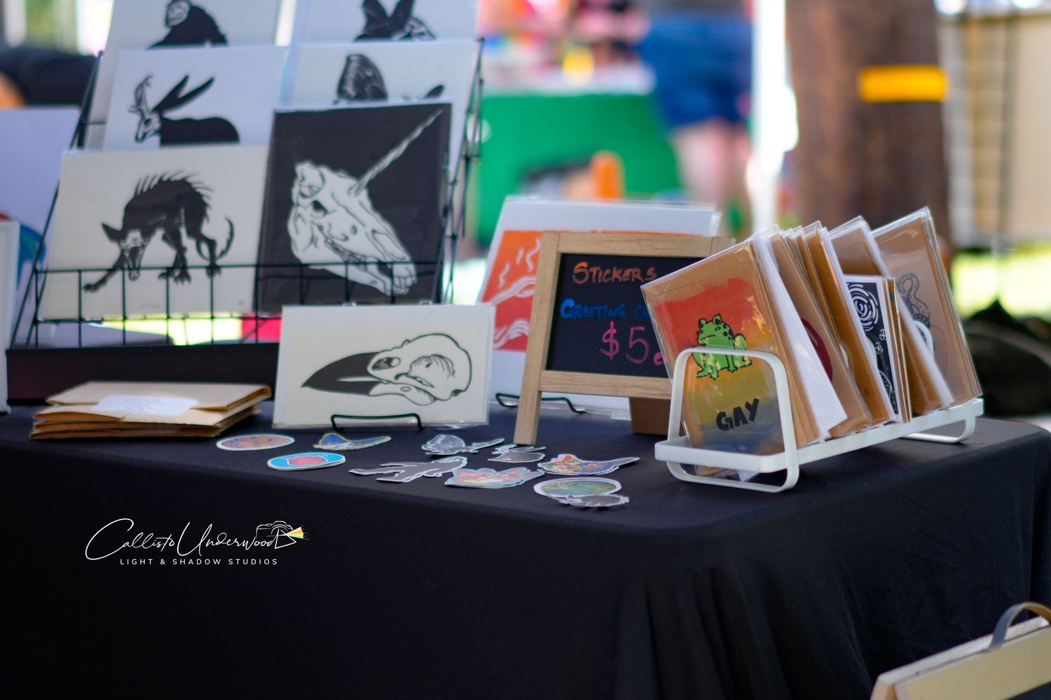 Handmade art prints, stickers and illustrated cards displayed on a black tablecloth at an outdoor artisan market stall.