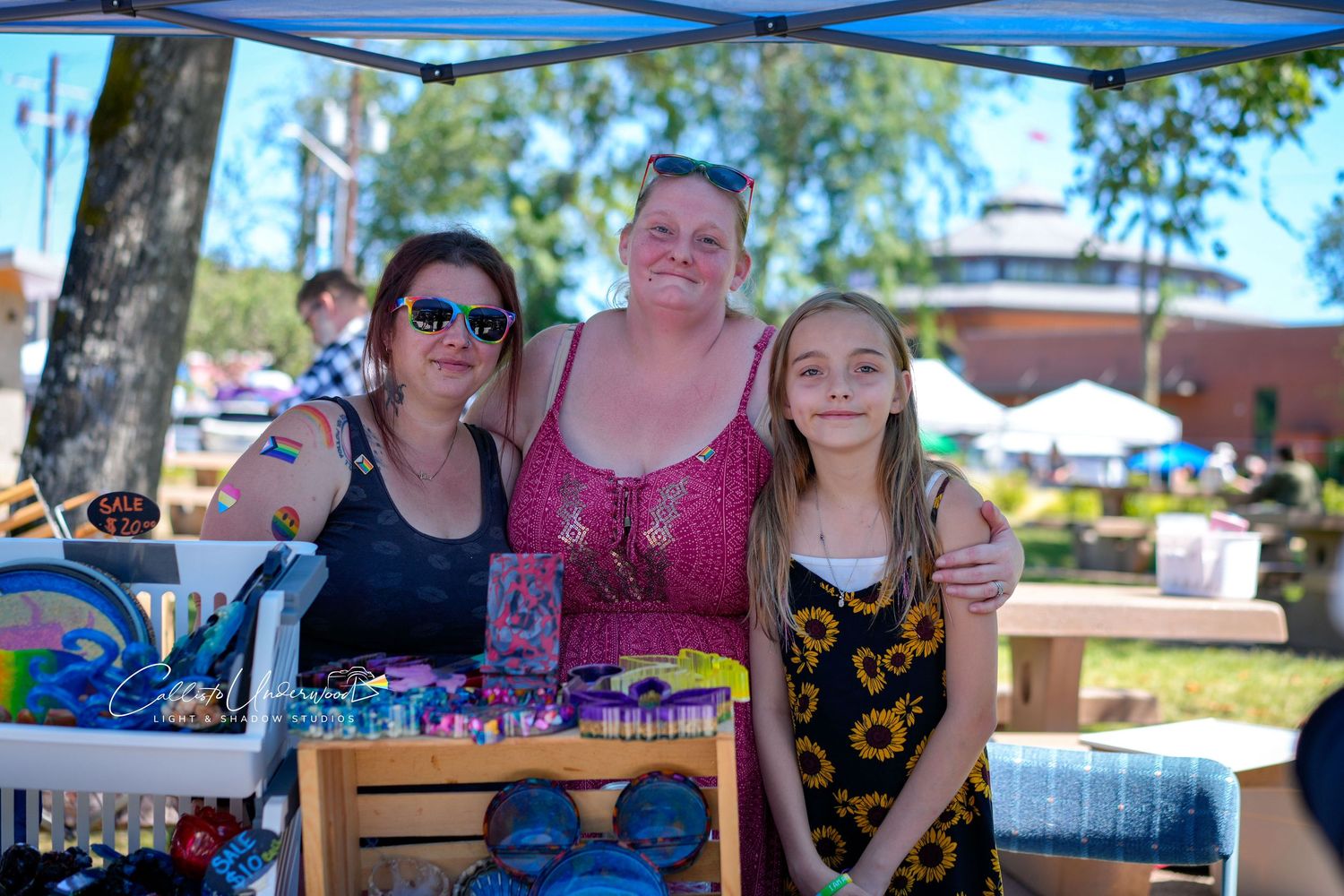 A woman, a girl and another vendor pose together behind a handmade crafts table at a sunny outdoor community market.