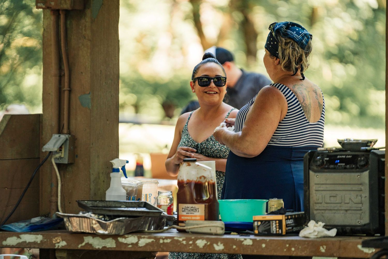 Two women smile and prepare food at a rustic outdoor serving table with sauces, trays and a portable cooler.