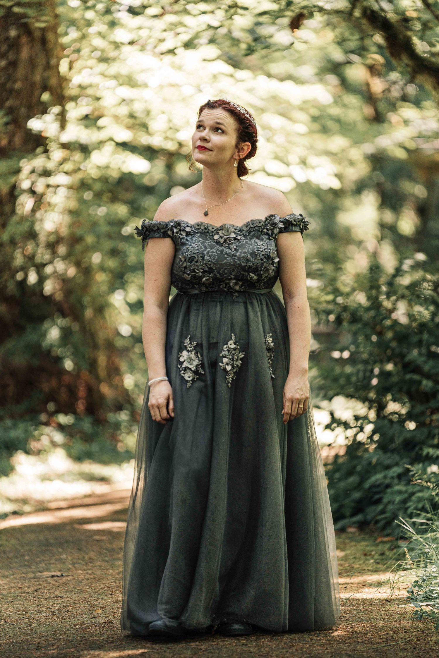 Woman in an elegant gray-green off-shoulder gown with floral embellishments gazes upward on a sunlit forest path.