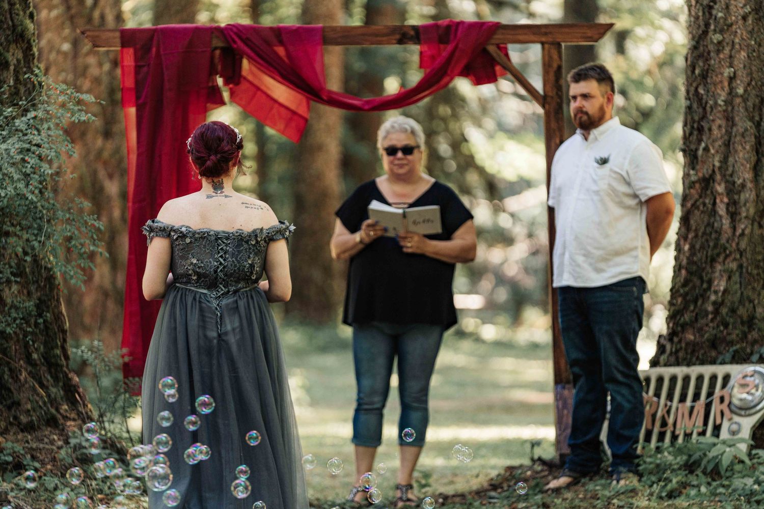A bride in a gray gown stands facing the officiant and groom beneath a red-draped arch while soap bubbles float nearby.