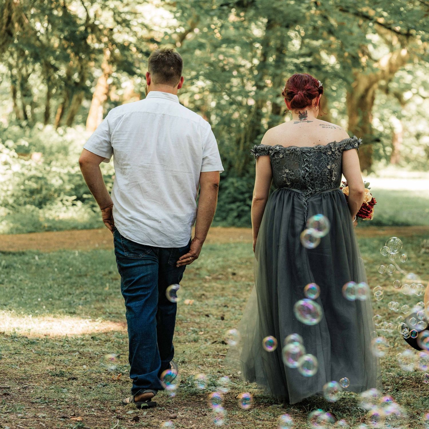 A newlywed couple walks away through a sunlit forest as soap bubbles float around them, the bride wearing a gray tulle gown.
