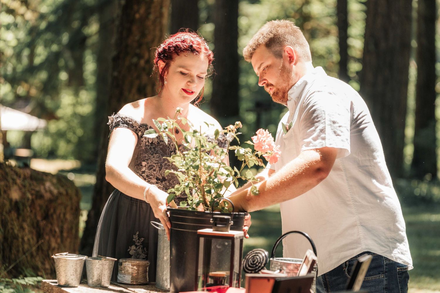 A smiling bride and groom tend a potted plant together at a wooden table during a forest wedding ritual.