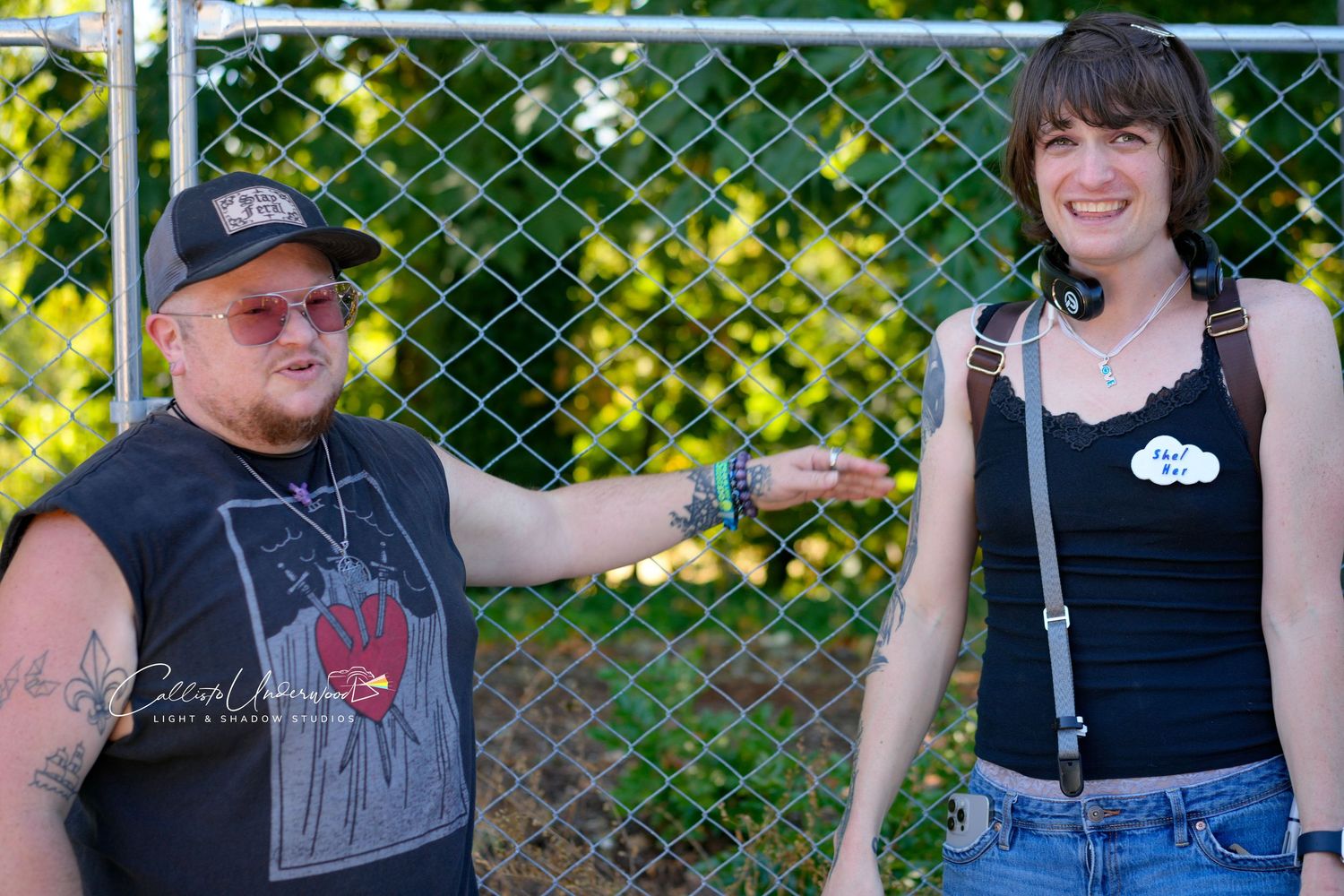 Smiling man in a baseball cap and sunglasses and a smiling woman with headphones pose near a chain-link fence at an outdoor event.