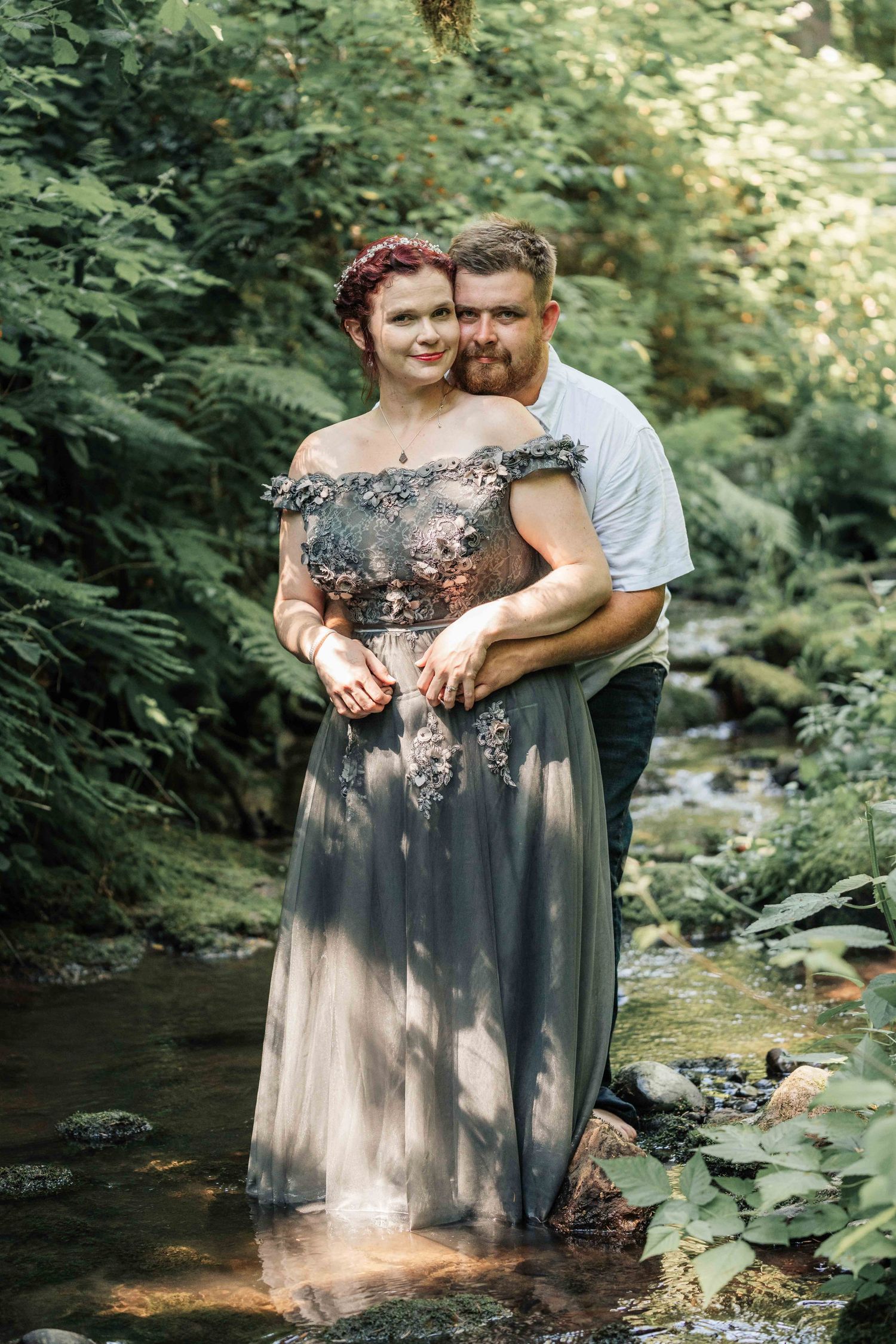 Smiling couple embracing in a gray tulle gown and white shirt while standing in a shallow forest stream surrounded by ferns.