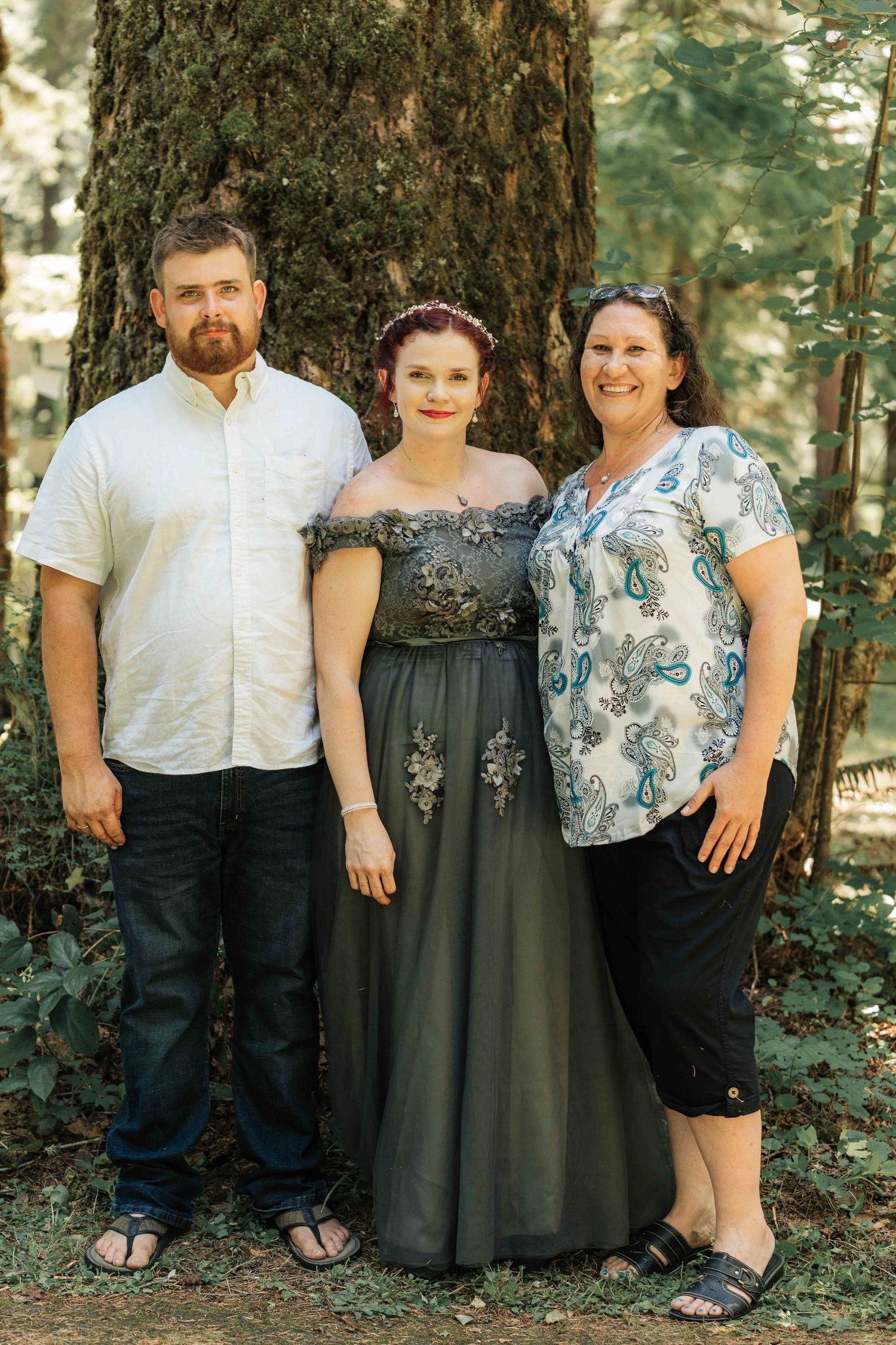 A bearded man, a bride in an off-shoulder gray dress, and a smiling woman in a floral top pose in front of a large mossy tree.