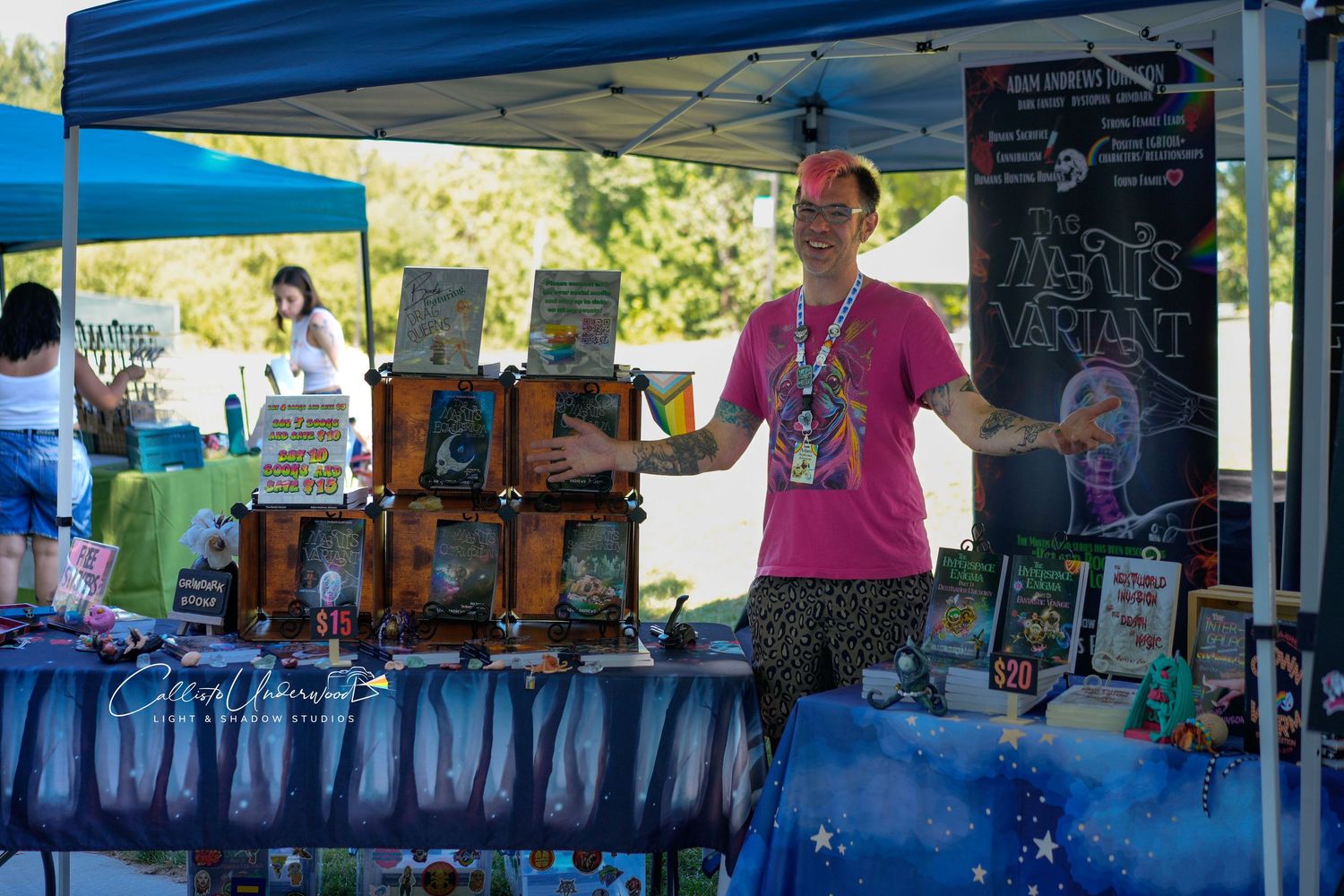 Author in a pink shirt gestures beside a vendor table of fantasy books and display boxes under a blue tent at an outdoor event.