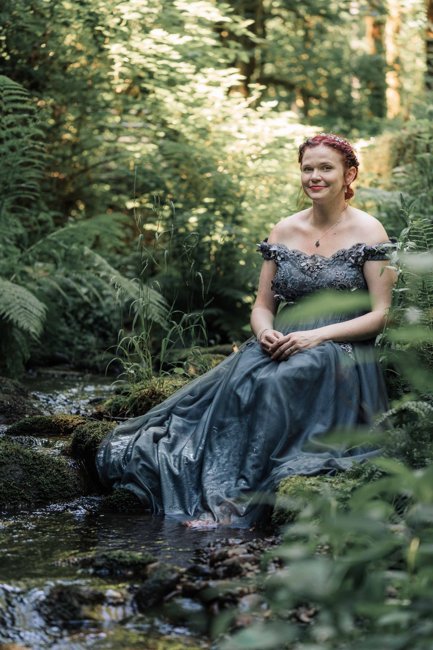 Red-haired woman in a gray lace-detailed gown sits on mossy rocks beside a forest stream, smiling softly among green ferns.