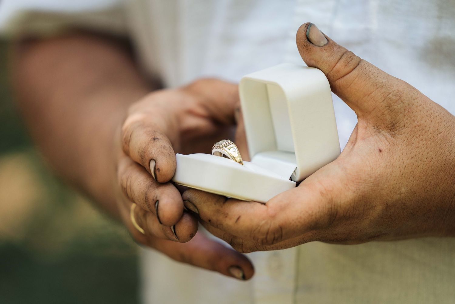 Close-up of a man's hands holding a white ring box with a gold ring inside at an outdoor wedding ceremony.