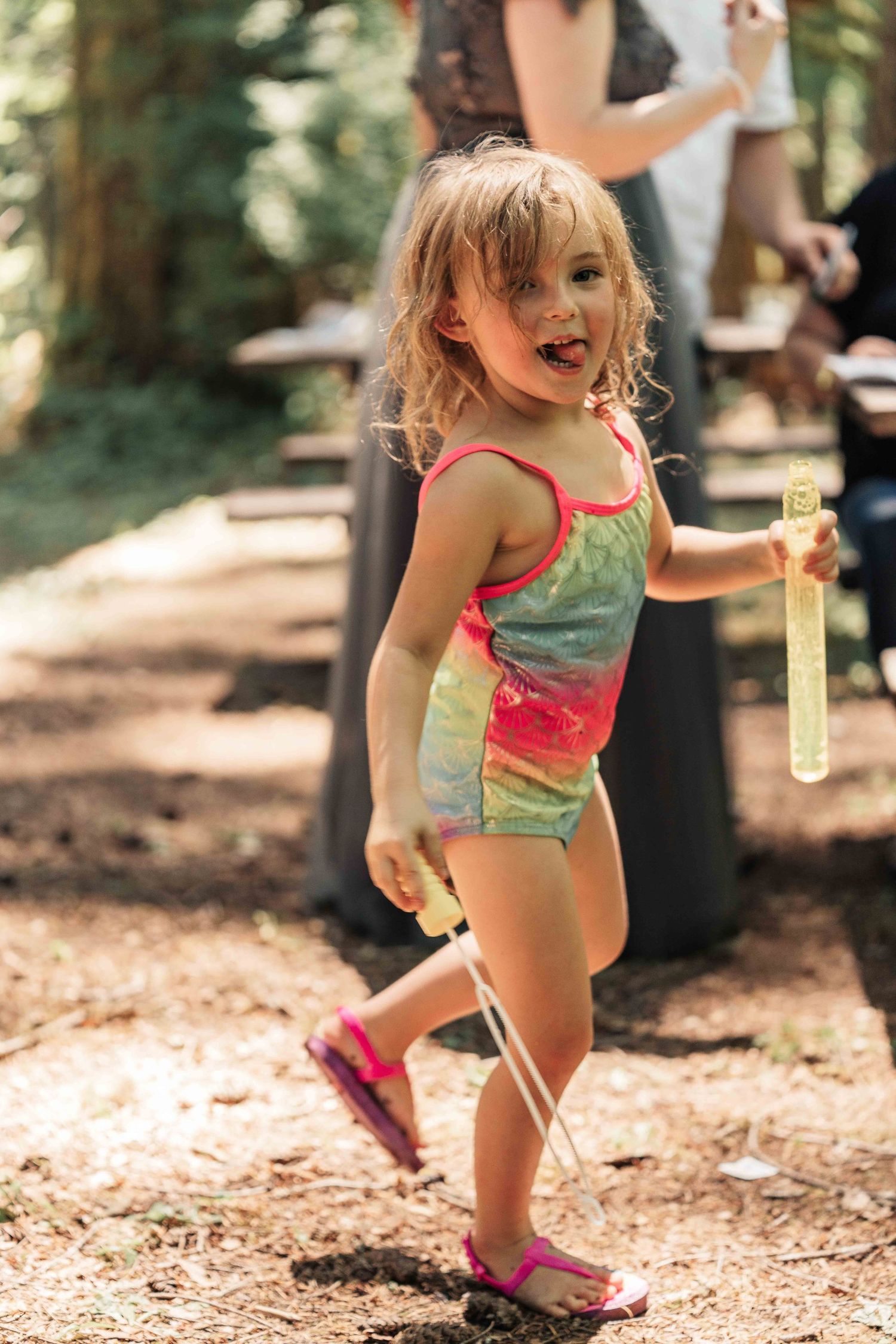 Playful little girl in a colorful swimsuit sticks out her tongue while holding a bubble wand and bottle on a wooded path.