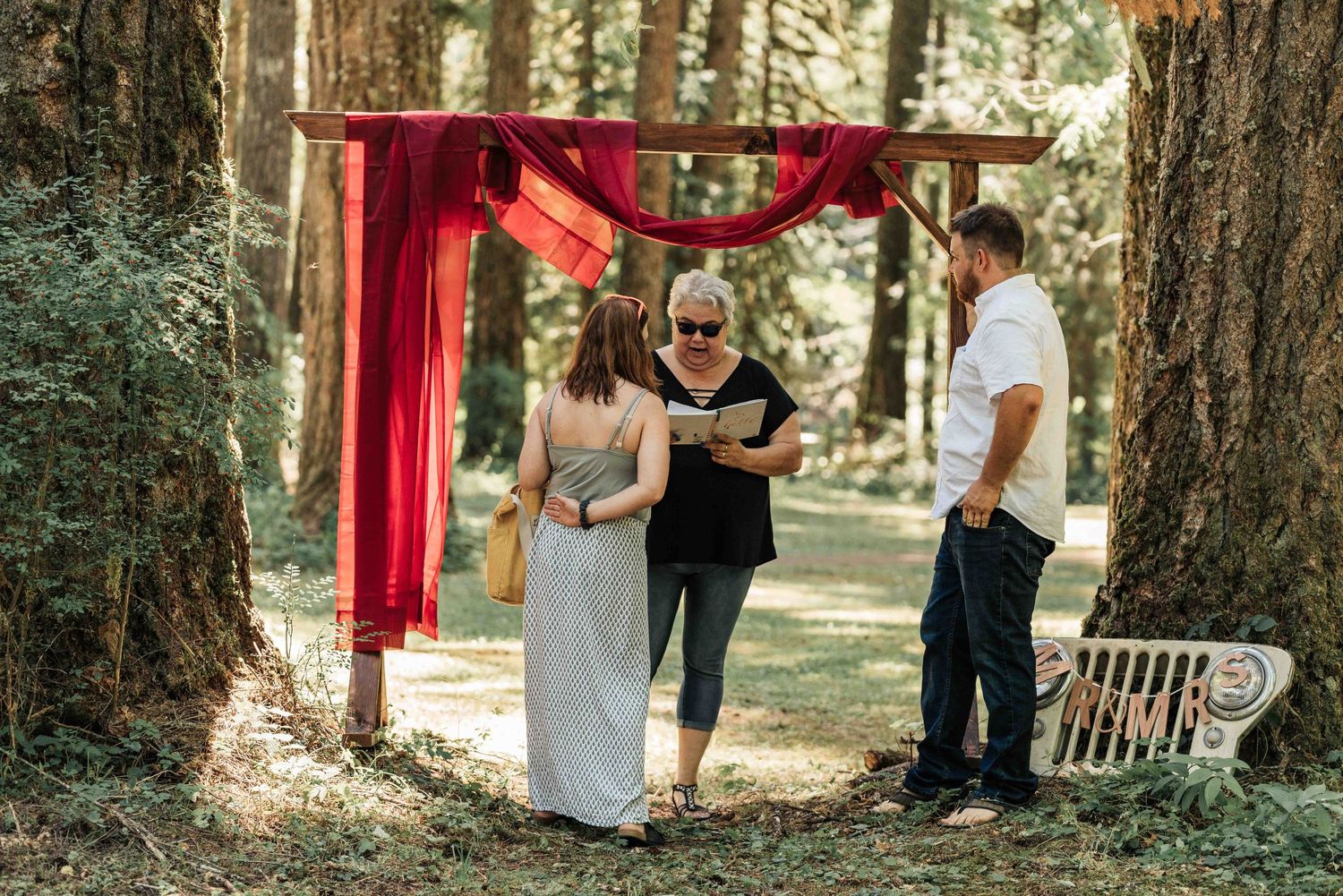 An officiant reads from a book under a red-draped wooden arch as a bride and groom stand among tall forest trees.