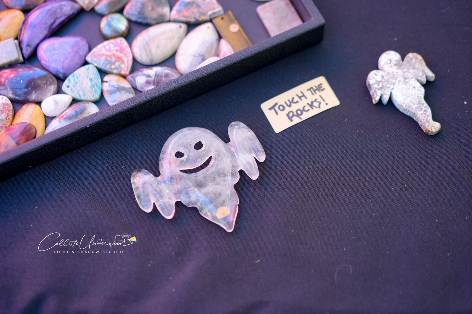 Smiling ghost-shaped stone carving and angel figurine beside a Touch the Rocks sign and polished gemstones on a market table.