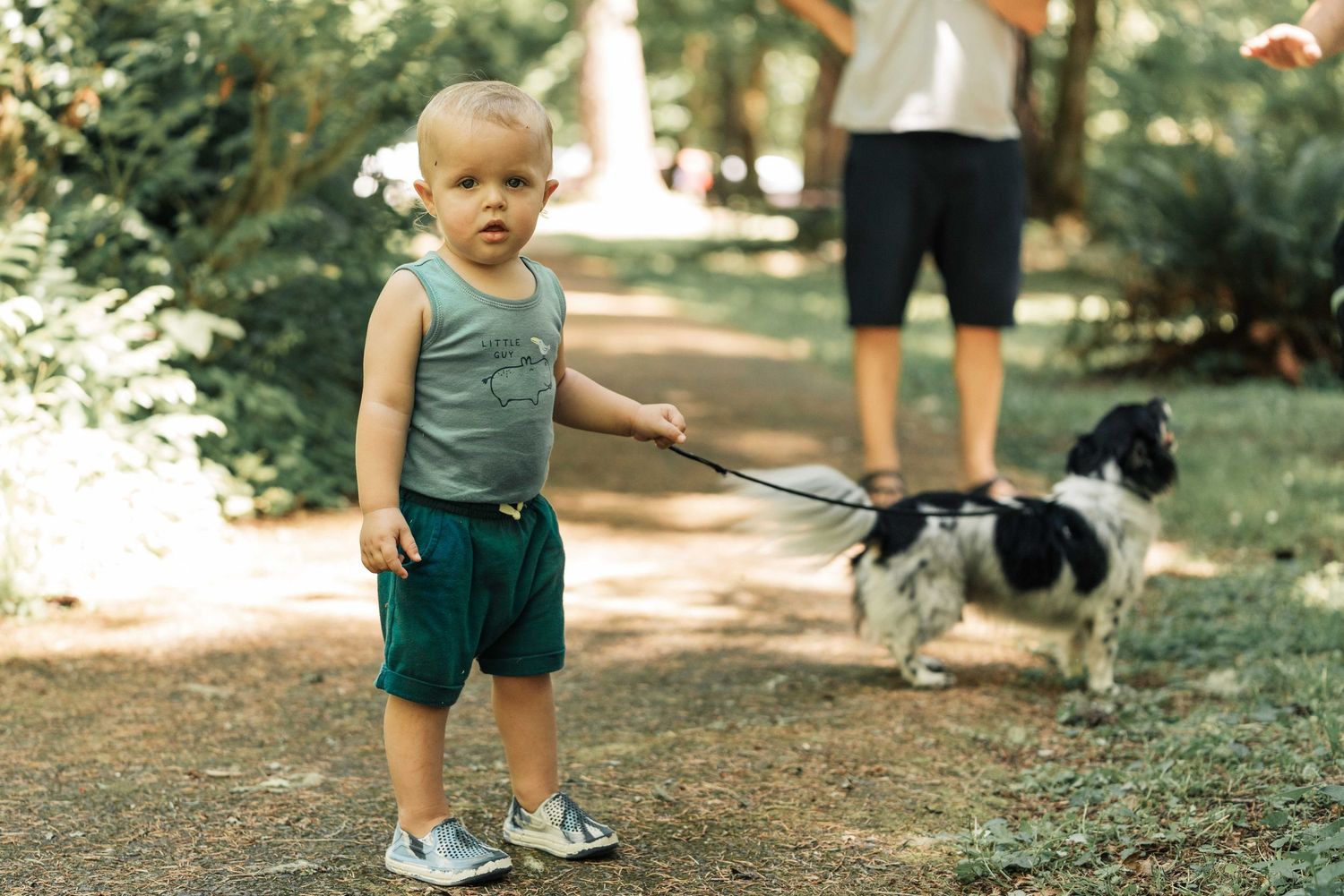 A toddler in a green tank top and shorts holds a leash attached to a small black and white dog on a forest path.